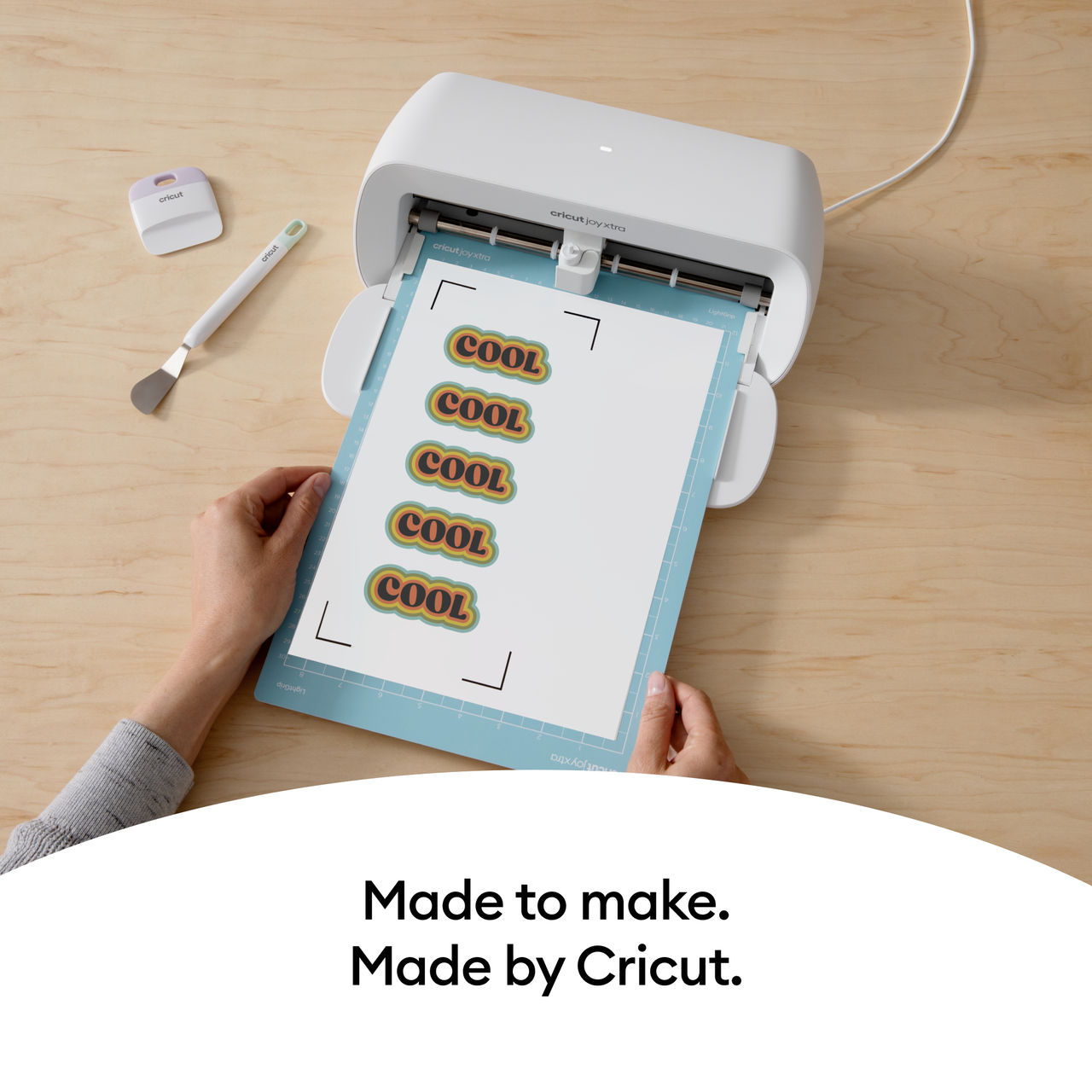 Eine weiße Cricut-Maschine schneidet COOL-Aufkleber auf einer blauen Matte. Hände halten die Matte.