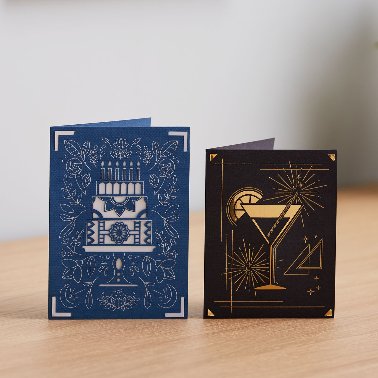 Zwei Grußkarten. Linke Karte: blau mit Kuchendesign. Rechte Karte: schwarz mit Cocktail-Design. Beide Karten liegen auf einer Holzoberfläche.