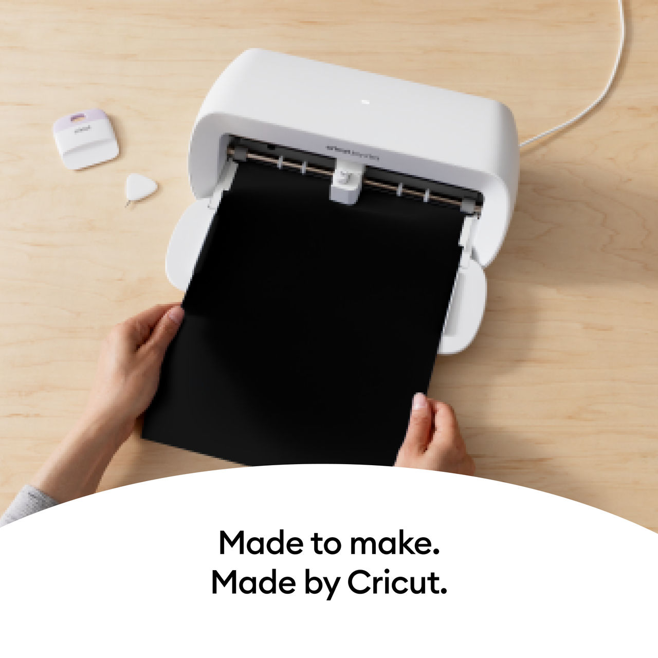 Eine Cricut-Maschine schneidet ein schwarzes Blatt, mit Text: 'Made to make. Made by Cricut.' und einem kleinen Cutter.
