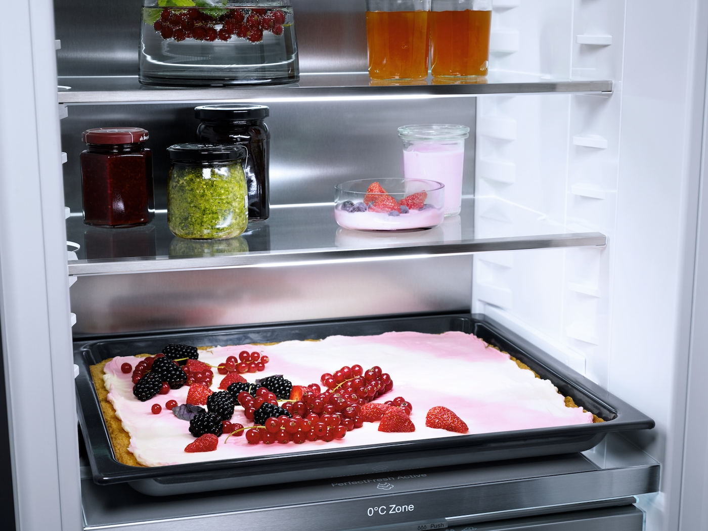 Ein Kühlschrankregal enthält Gläser mit Marmelade, Pesto, Joghurt und eine große Schale mit Beeren auf einem Kuchen.