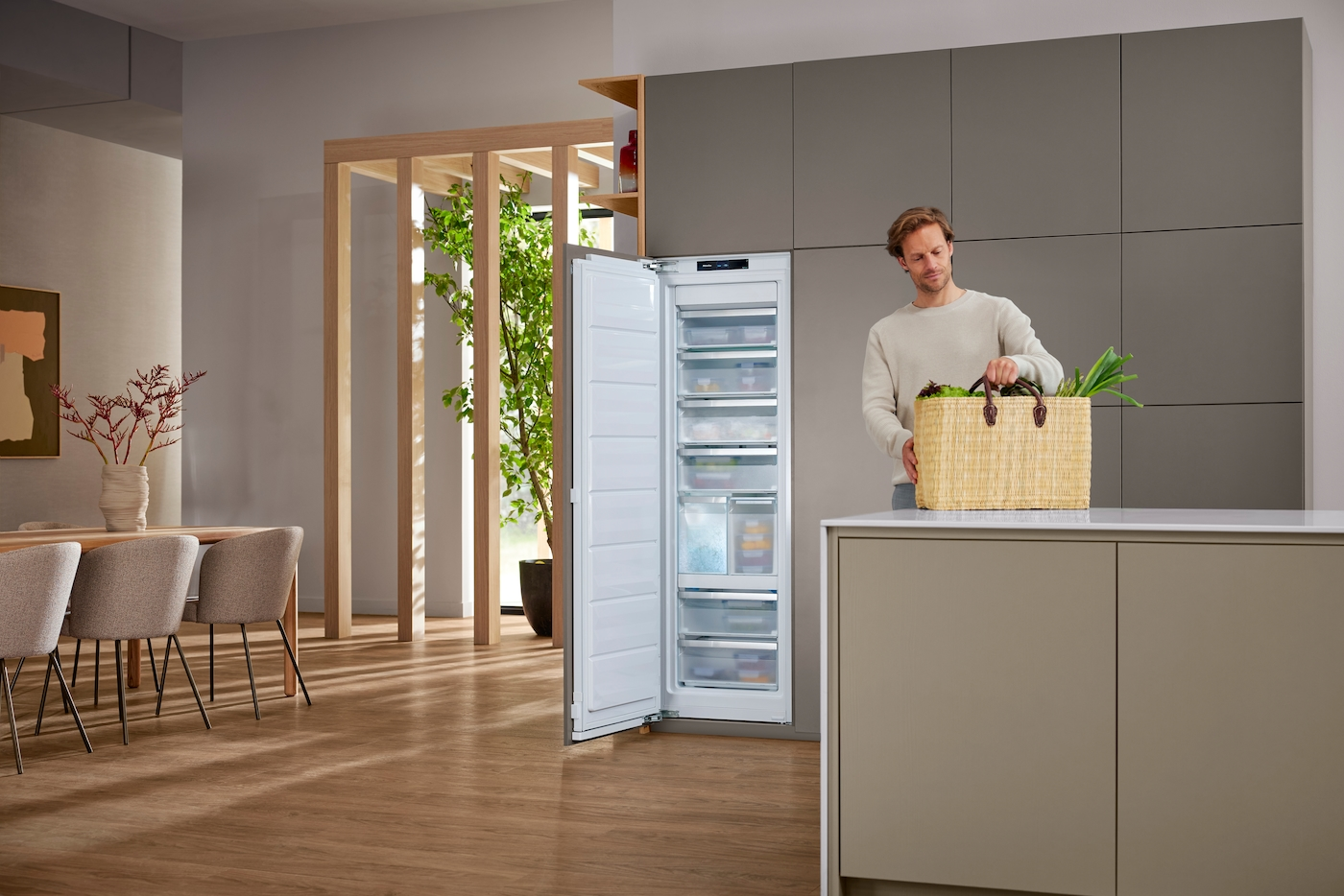Mann mit Einkaufskorb voller Gemüse vor einem offenen Kühlschrank in einer modernen Küche.