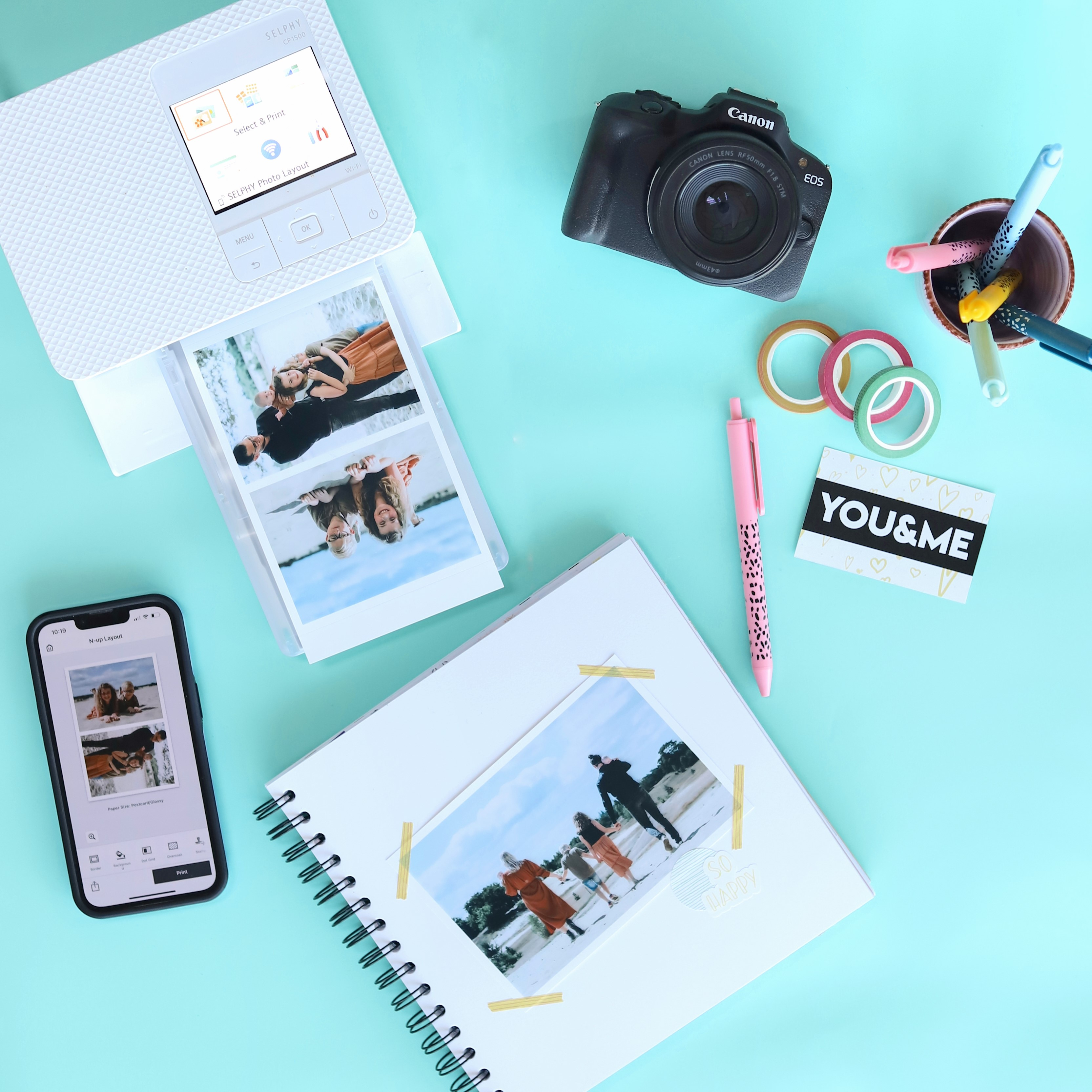 A photo printing setup. Printer, camera, phone, journal, pens, washi tape, photos. Light blue surface.