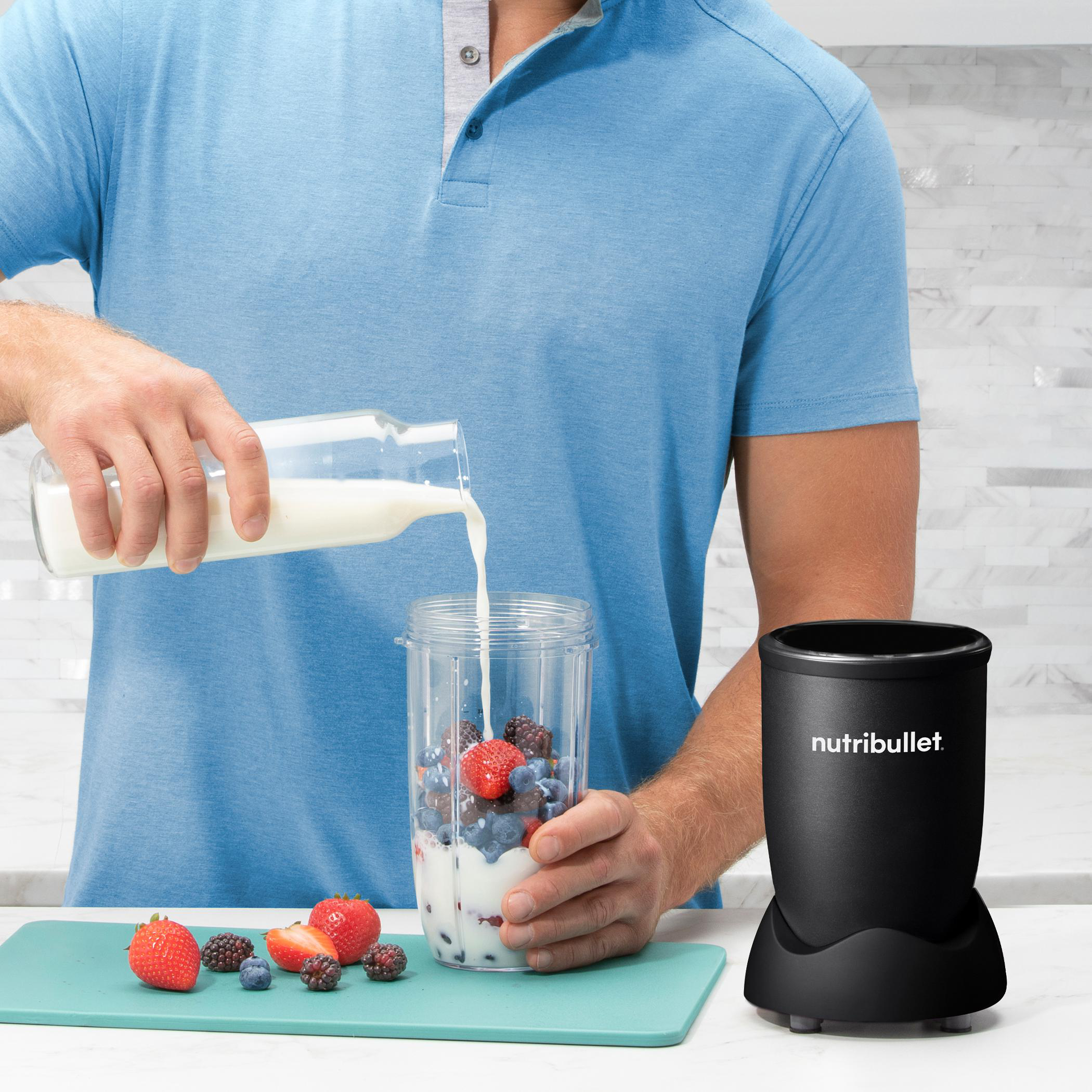 Eine Person gießt Milch in einen Mixer mit Früchten. Ein Mixer daneben.
