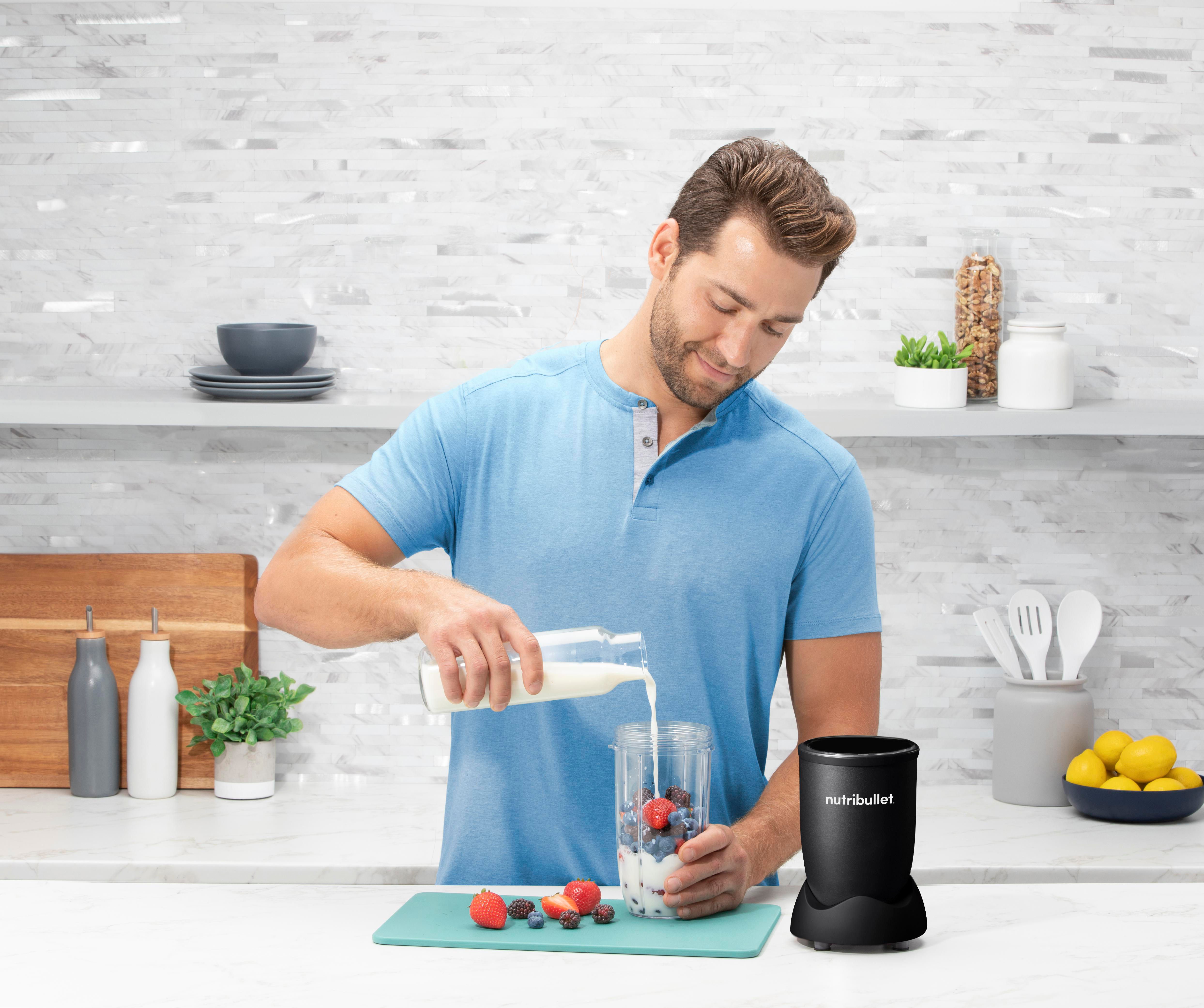 Mann gießt Milch in einen Mixerbecher mit Obst, weiße Küche.