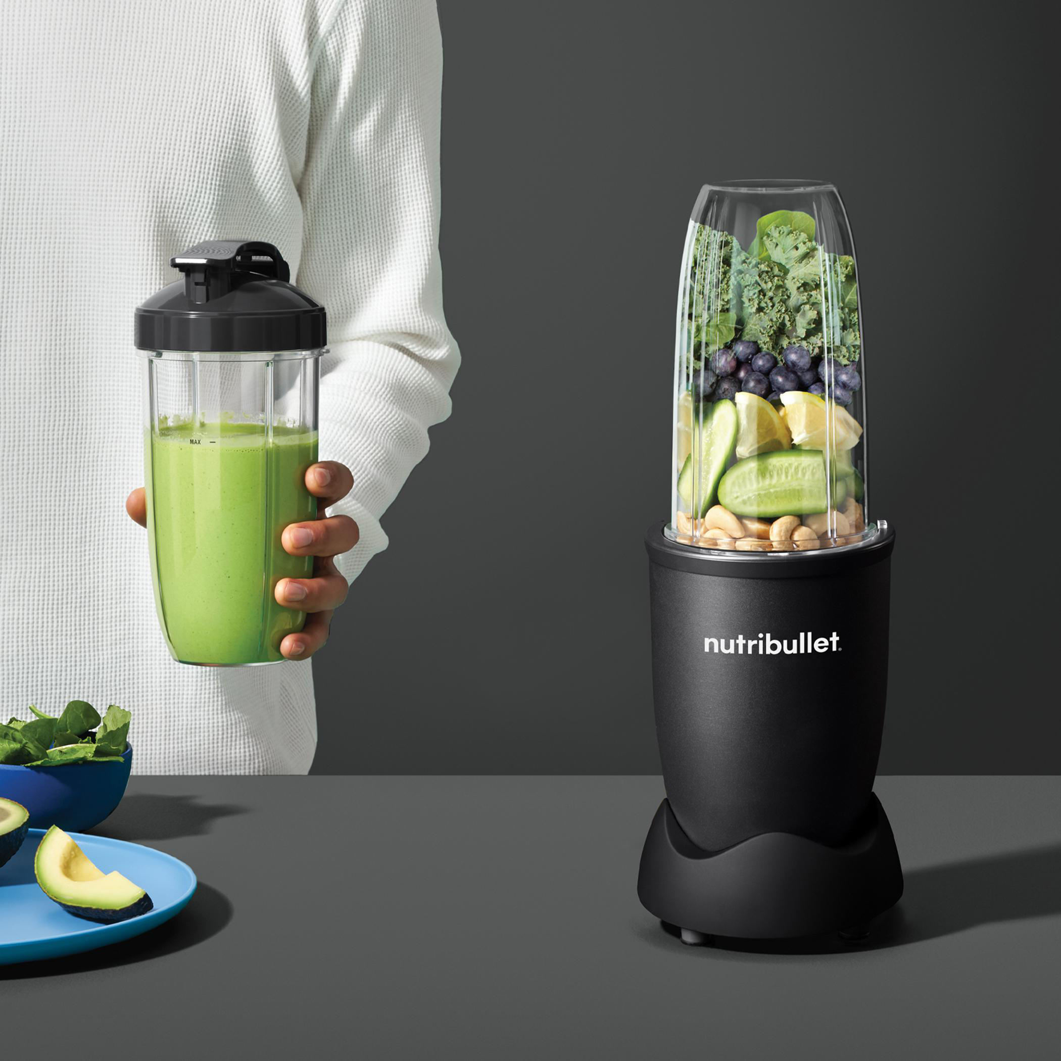 Eine Person hält einen grünen Smoothie in einem Nutribullet. Mixer mit Zutaten. Grauer Hintergrund.