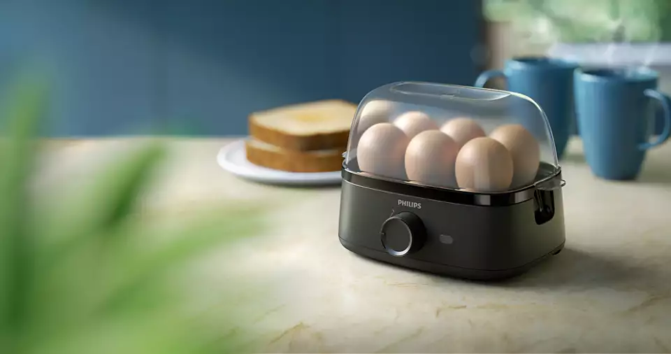 Eierkocher mit Eiern darin. Schwarze Farbe, transparente Abdeckung, Marke Philips. Toast und Tassen im Hintergrund.