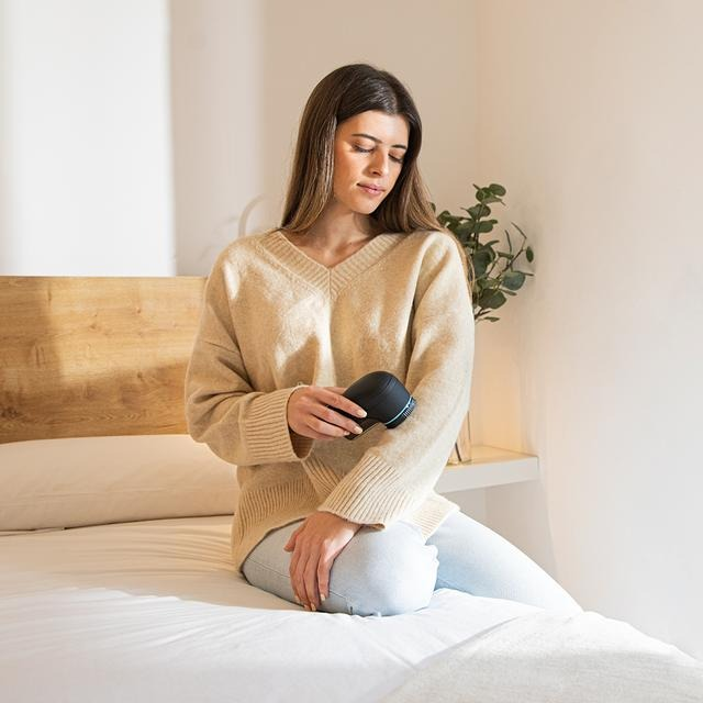 Mujer sentada en una cama, sosteniendo un dispositivo negro y mirándolo.
