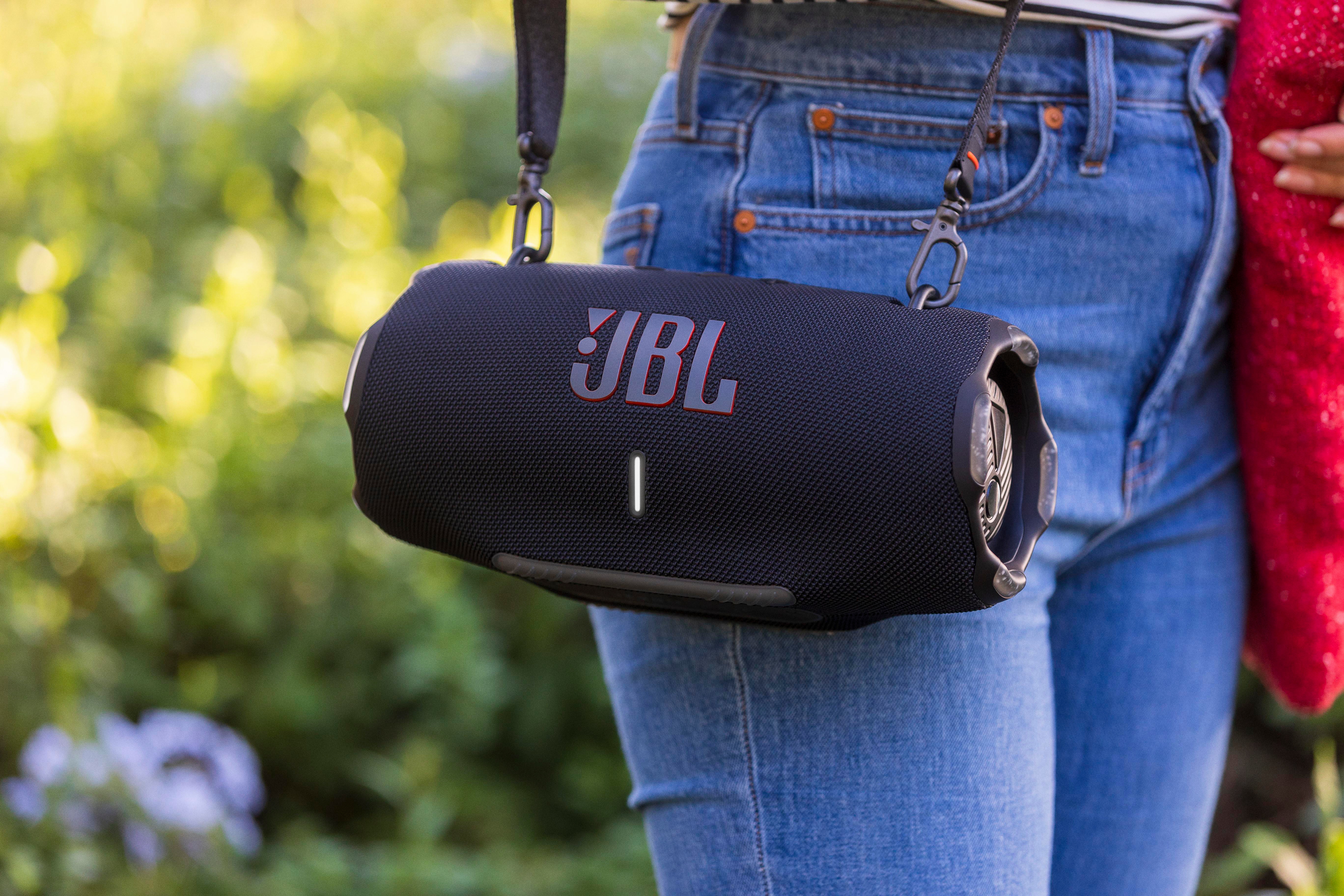 Ein schwarzer JBL-Lautsprecher mit Riemen, getragen von einer Person in Jeans. Der Hintergrund ist verschwommenes Grün.