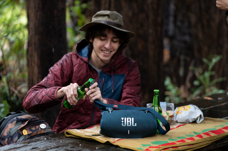 Ein Mann öffnet draußen eine Bierflasche mit einem JBL-Lautsprecher und Picknick-Setup.