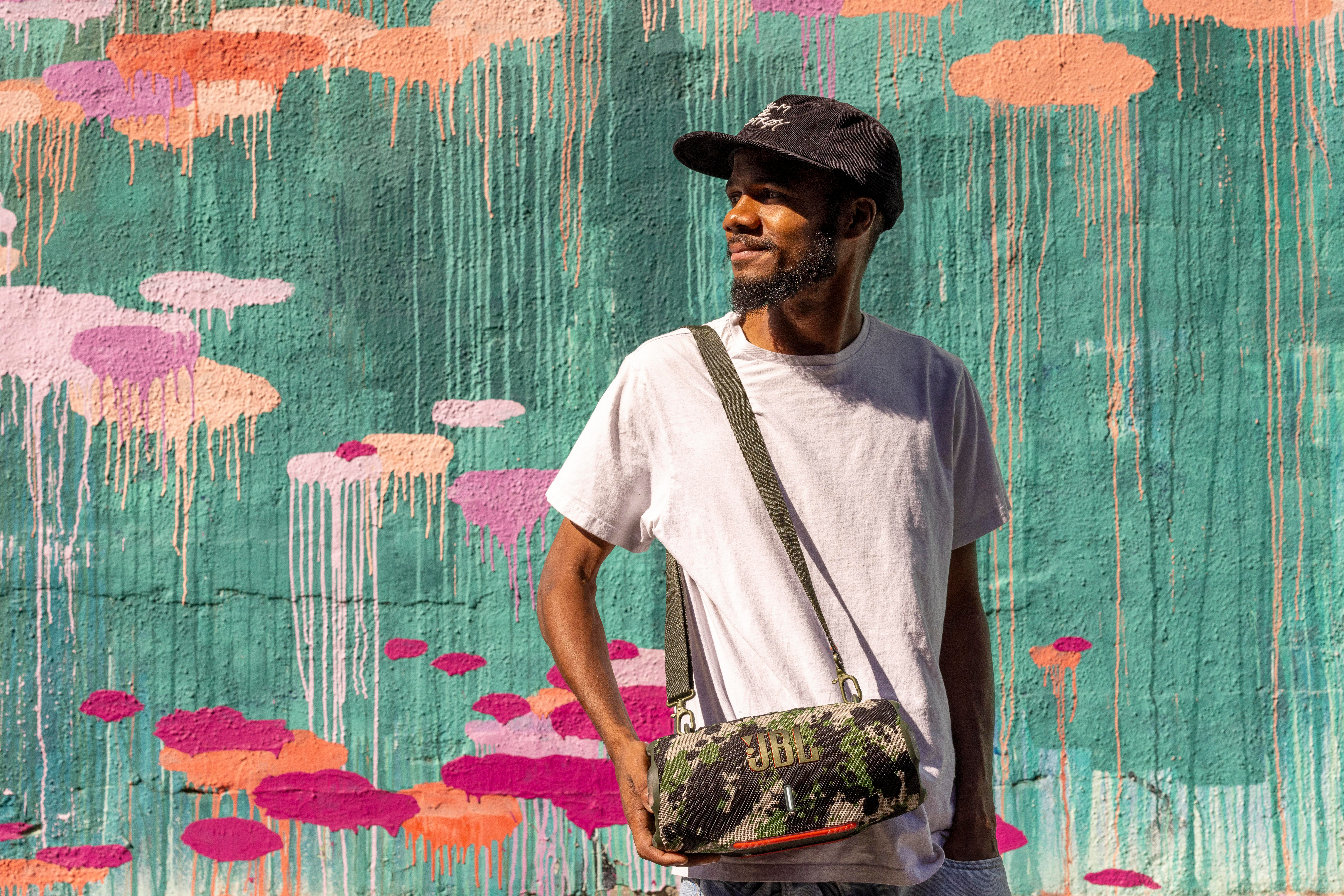 Mann mit Hut und Lautsprechertasche vor einer Graffiti-Wand.