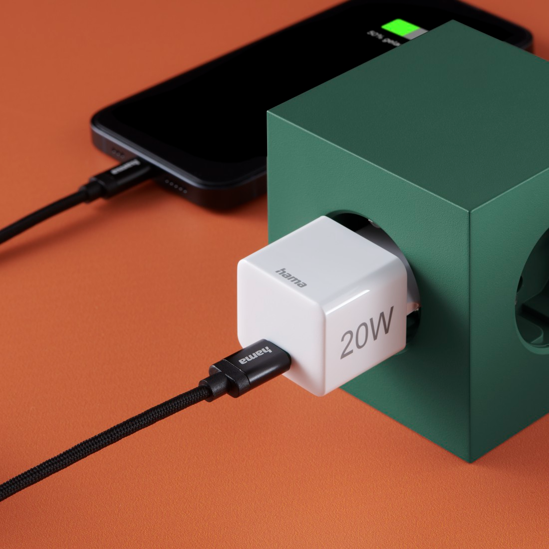 Egy fehér 20W-os Hama töltő egy zöld kockába van dugva, mellette egy fekete telefon.