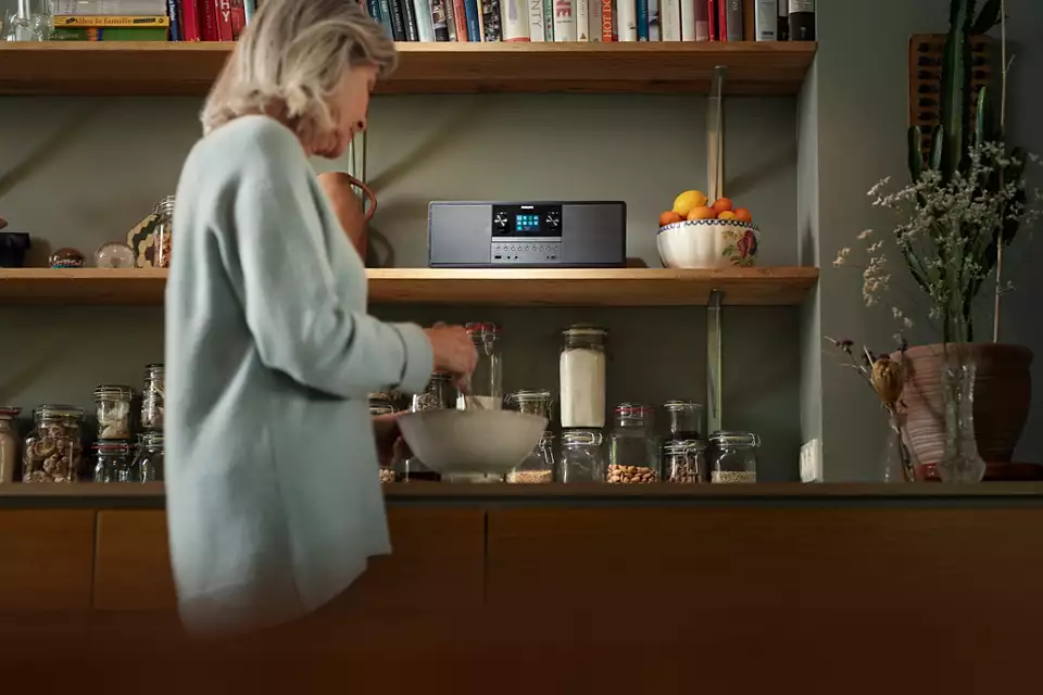 Una donna in cucina mescola in una ciotola. Gli scaffali contengono barattoli, libri e uno stereo. Una ciotola di frutta e una pianta sono visibili.