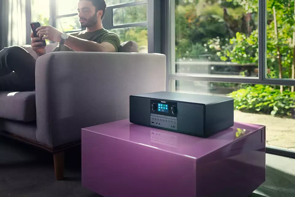 Un uomo usa un telefono su un divano. Un impianto stereo è su un tavolo rosa. Fuori c'è del fogliame verde.