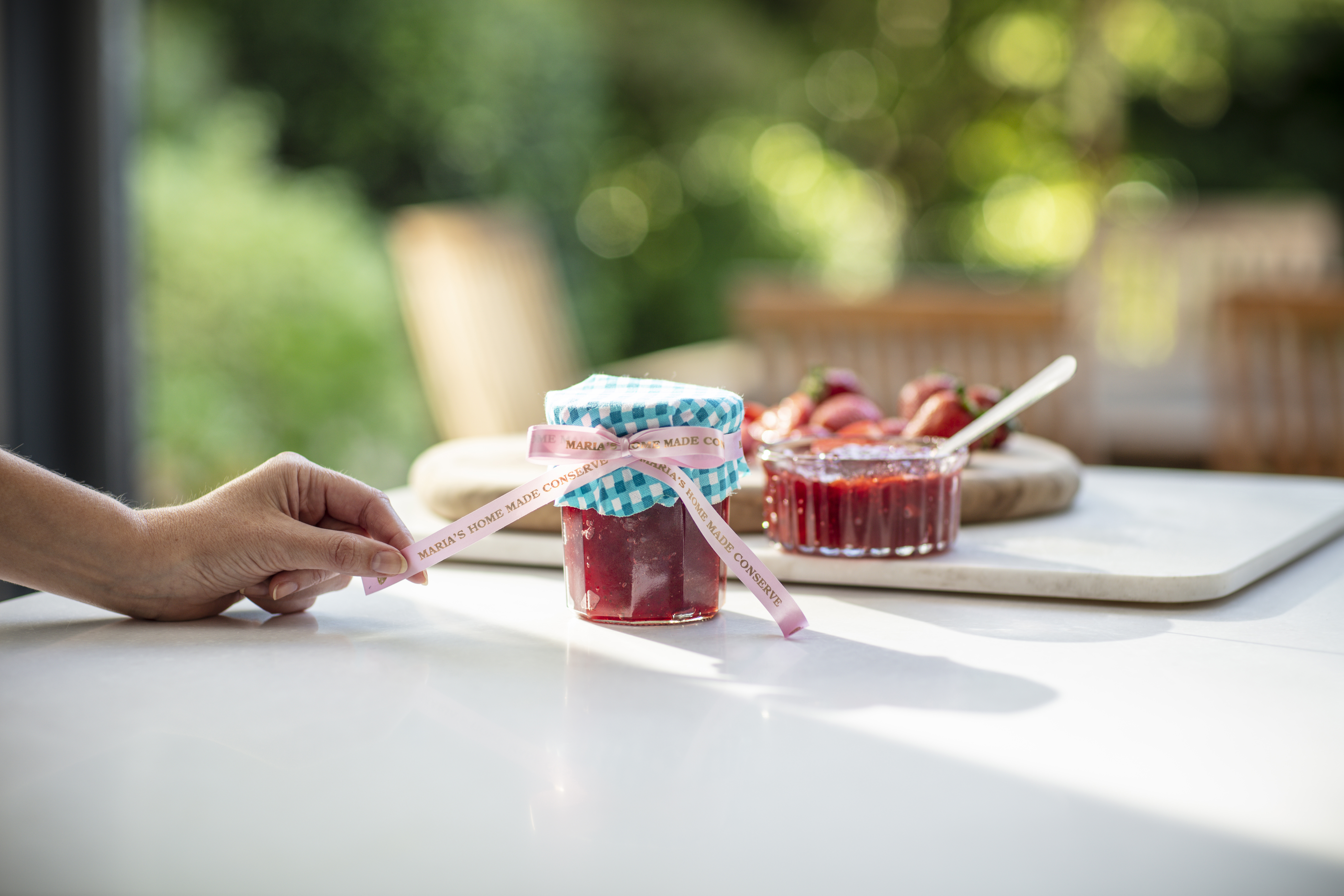 Hand hält Band um ein Marmeladenglas auf einem Tisch. Erdbeeren und Marmelade auf Schneidebrett.