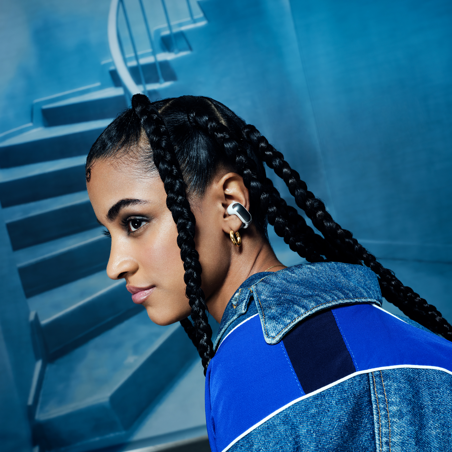 A woman with braids wearing a denim jacket and earphones. Blue stairs in the background.