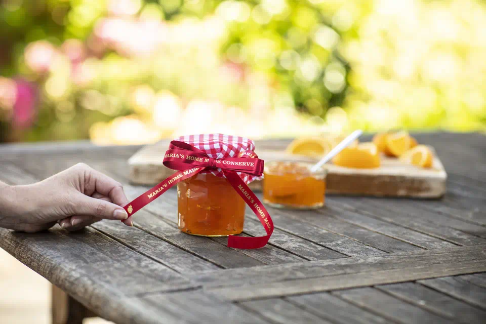 Barattolo di marmellata con nastro rosso e fette d'arancia su un tavolo di legno al sole.