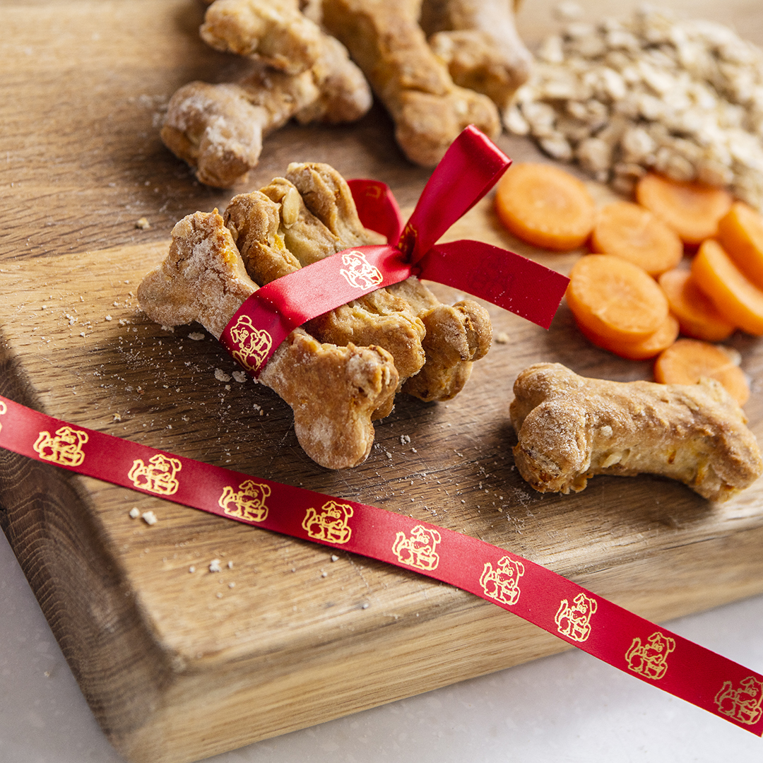 Tavola di legno con biscotti per cani a forma di osso e un nastro rosso, più carote tagliate e avena.