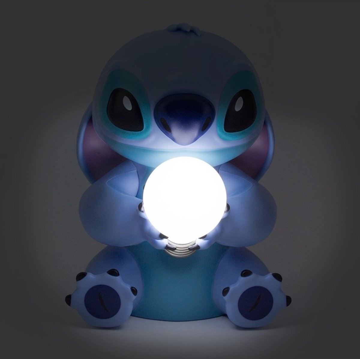 A blue Stitch lamp holds a glowing white orb in its hands, in a dark room.