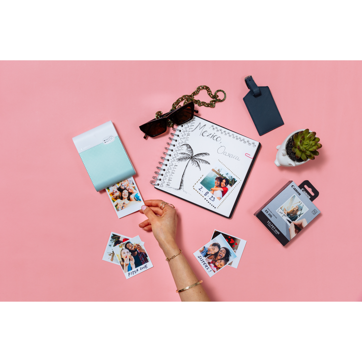 Photos, a printer, sunglasses, and a notebook are on a pink surface. A hand is holding a photo.
