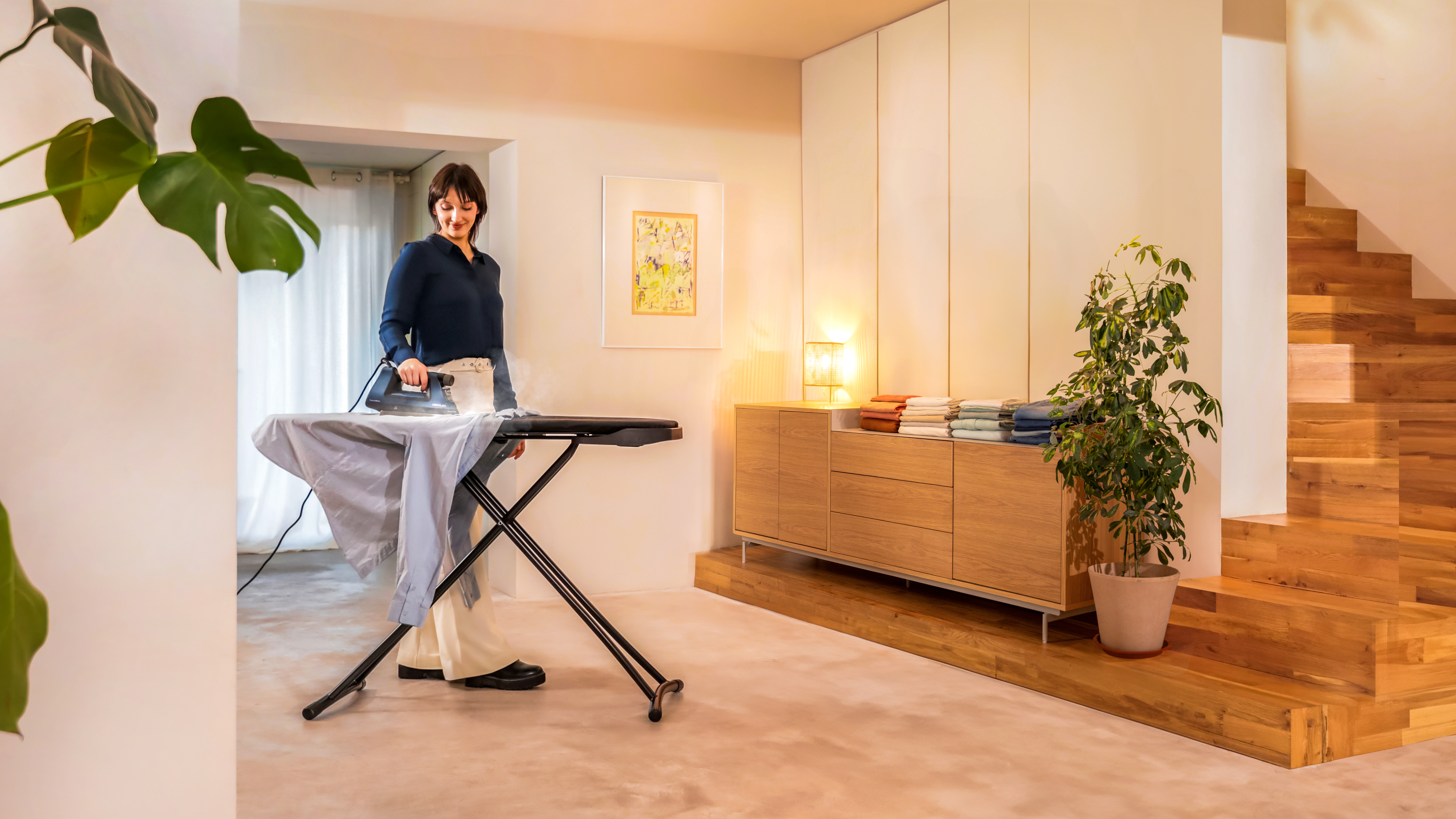 Mujer planchando camisa en tabla de planchar. Fondo: muebles y escaleras.