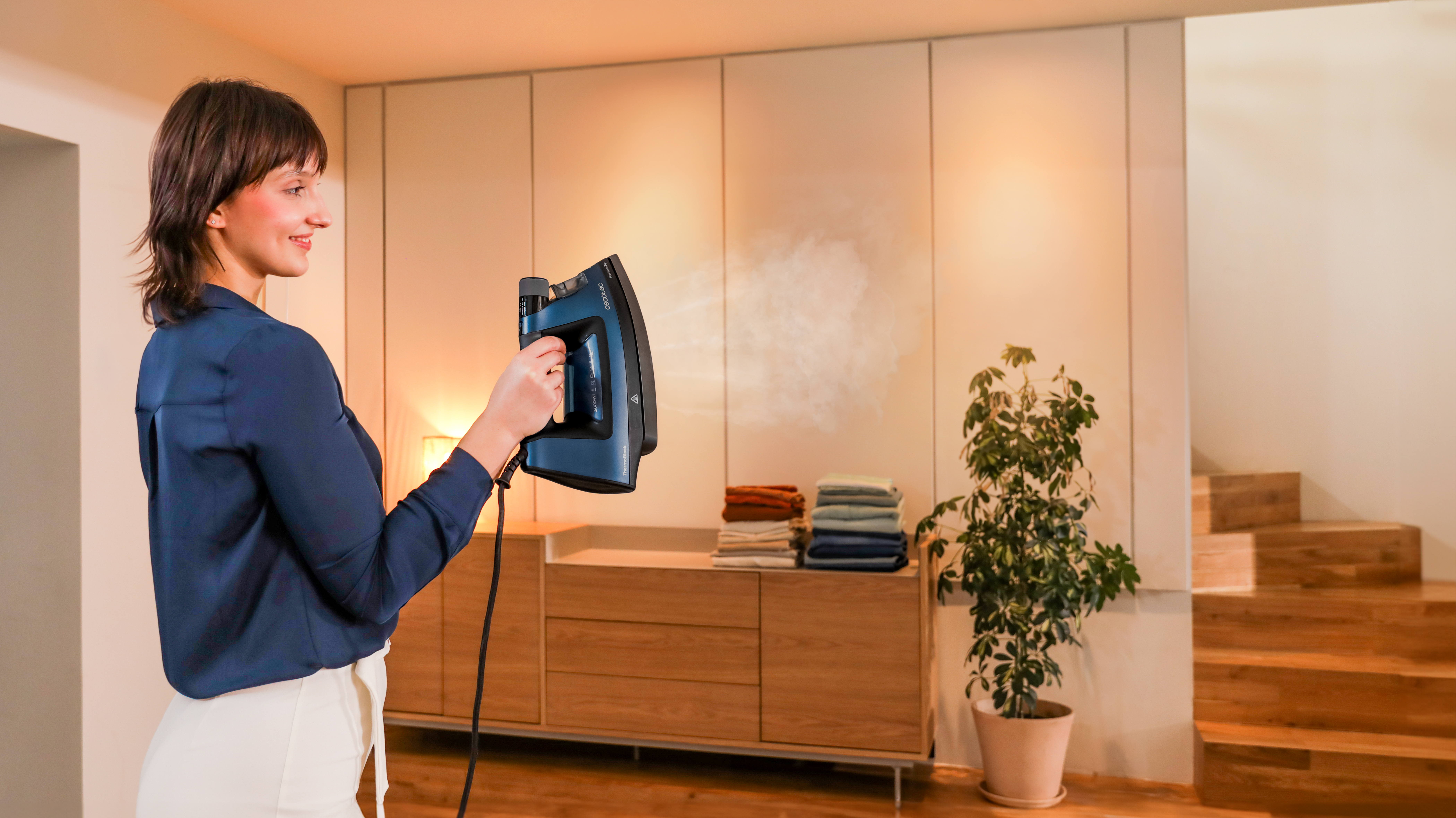Una mujer planchando ropa con una plancha de vapor. En el fondo, hay un mueble de madera y escaleras.