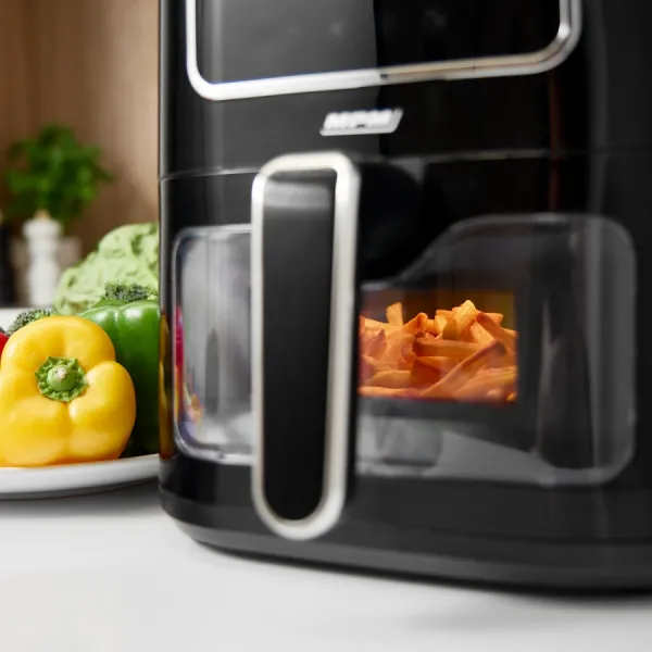 Czarna frytkownica powietrzna z przezroczystym oknem pokazującym frytki w środku; warzywa w tle.