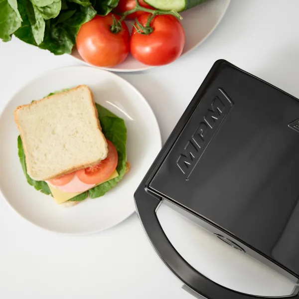 Kanapka na talerzu, pomidory i sałata na białej powierzchni obok czarnego opiekacza do kanapek.