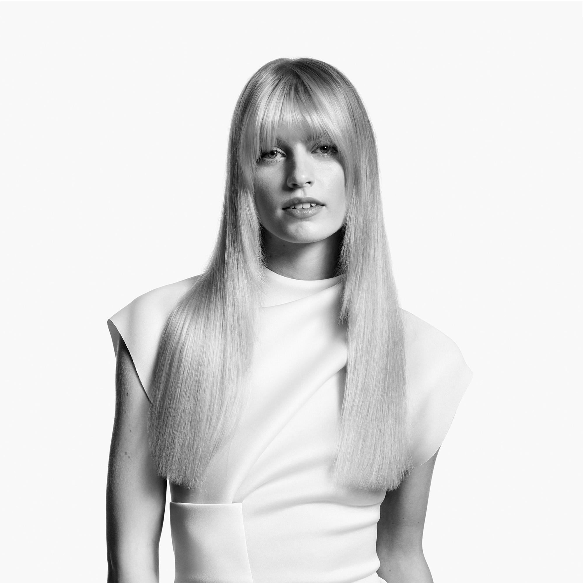 Blonde Frau mit glattem Haar und Pony, trägt ein weißes Oberteil, vor weißem Hintergrund.