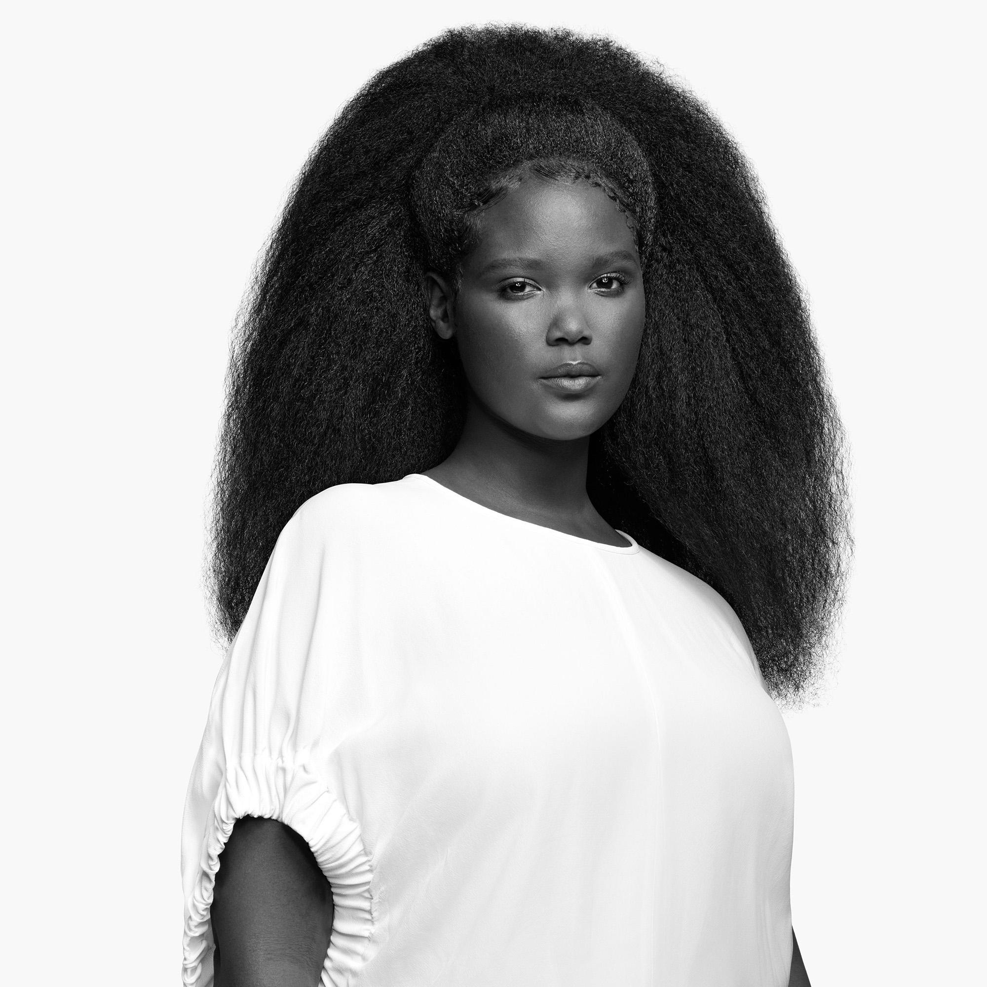 Eine Frau mit voluminösem, schwarzem Haar, in einem weißen Oberteil, blickt direkt in die Kamera.