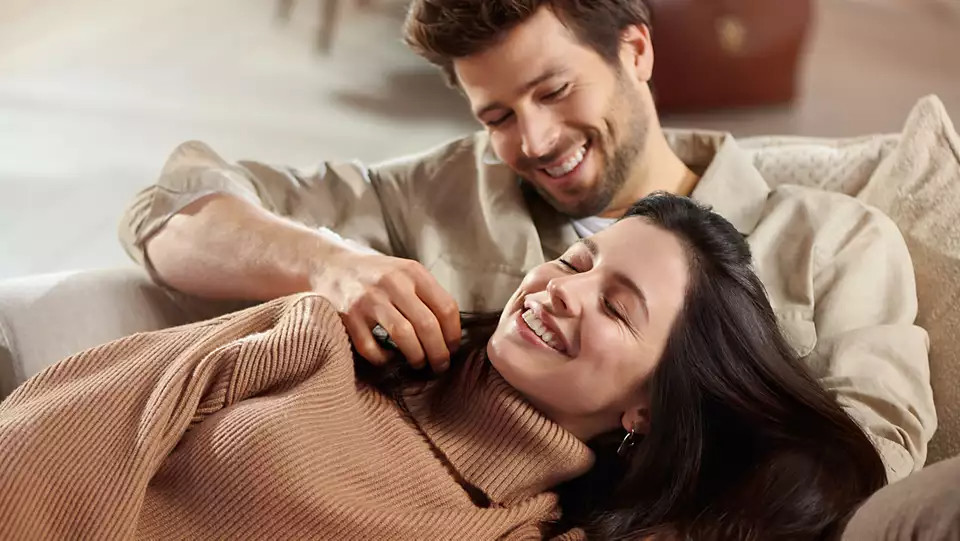 Een man en een vrouw lachen op een bank, de man raakt de nek van de vrouw aan.