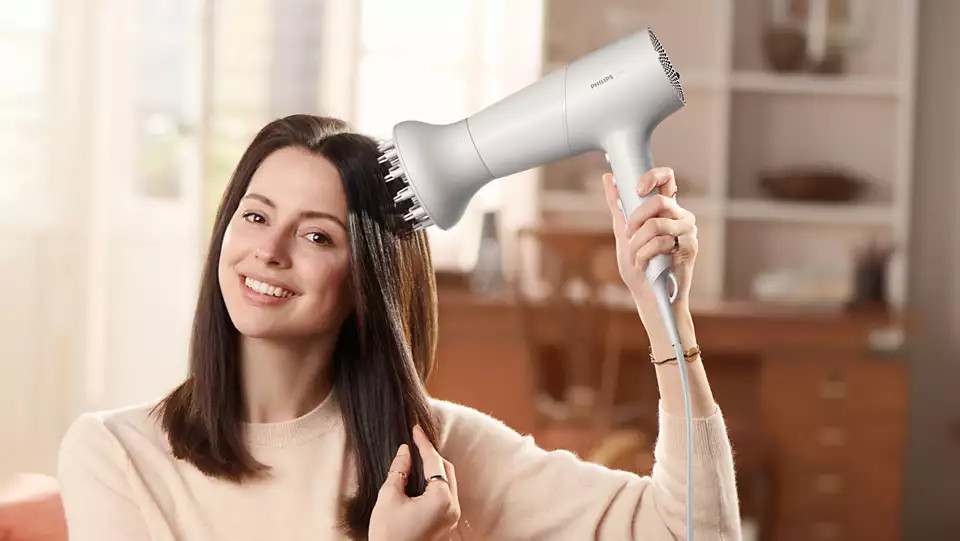 Een vrouw glimlacht terwijl ze een grijze föhn gebruikt, haar donkere haar droogt, met een huiselijke achtergrond.