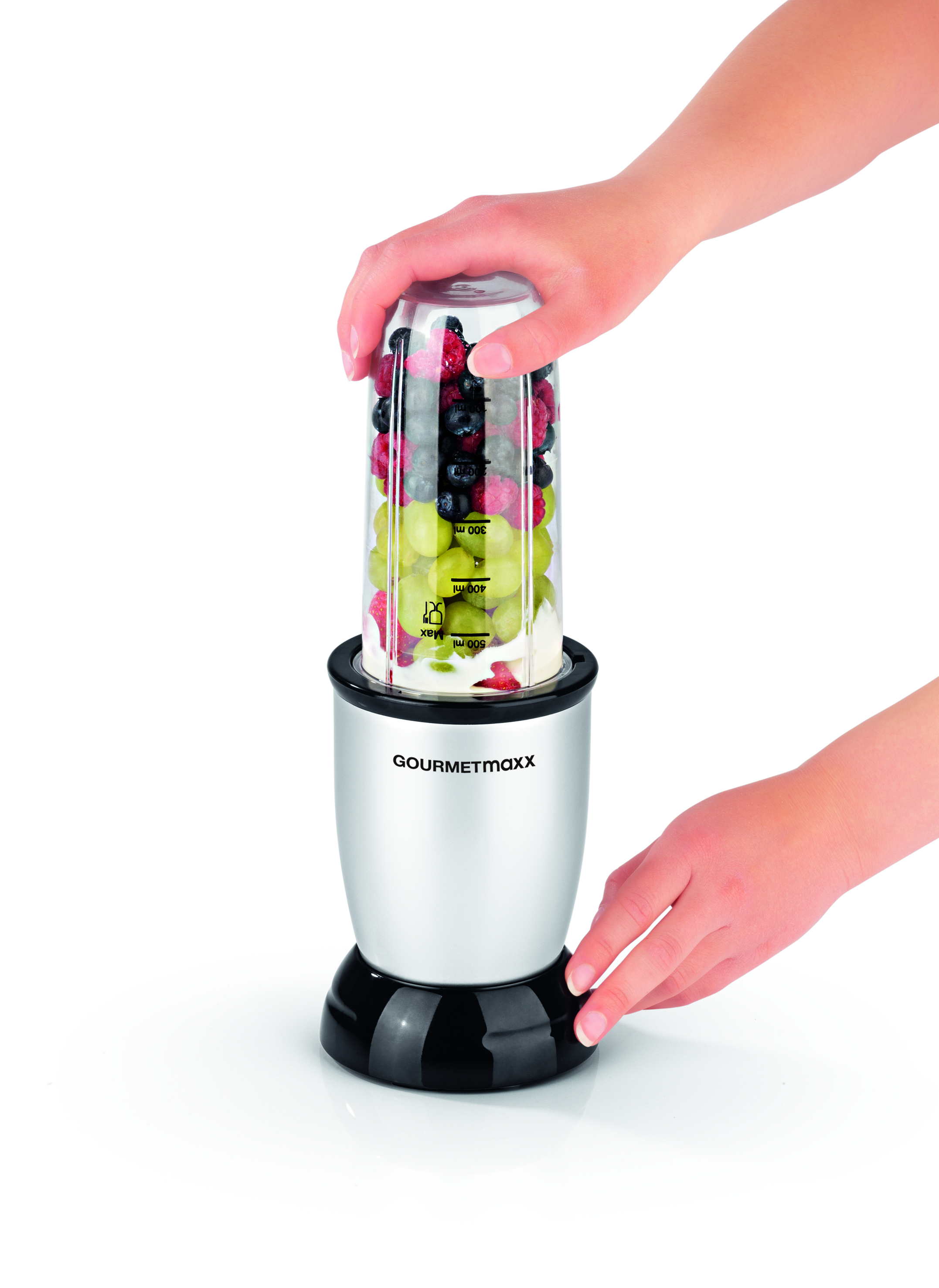 Hände setzen einen mit Früchten gefüllten Mixbecher auf einen Sockel, Früchte sind sichtbar.