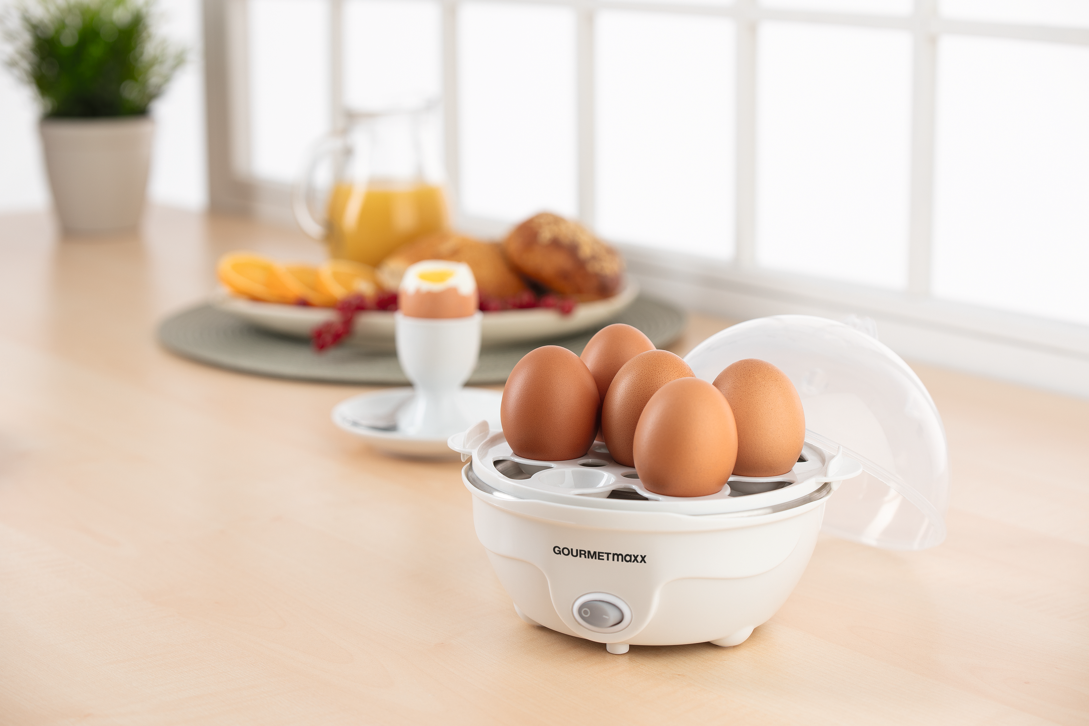Eierkocher mit 4 Eiern, Frühstücksessen im Hintergrund.