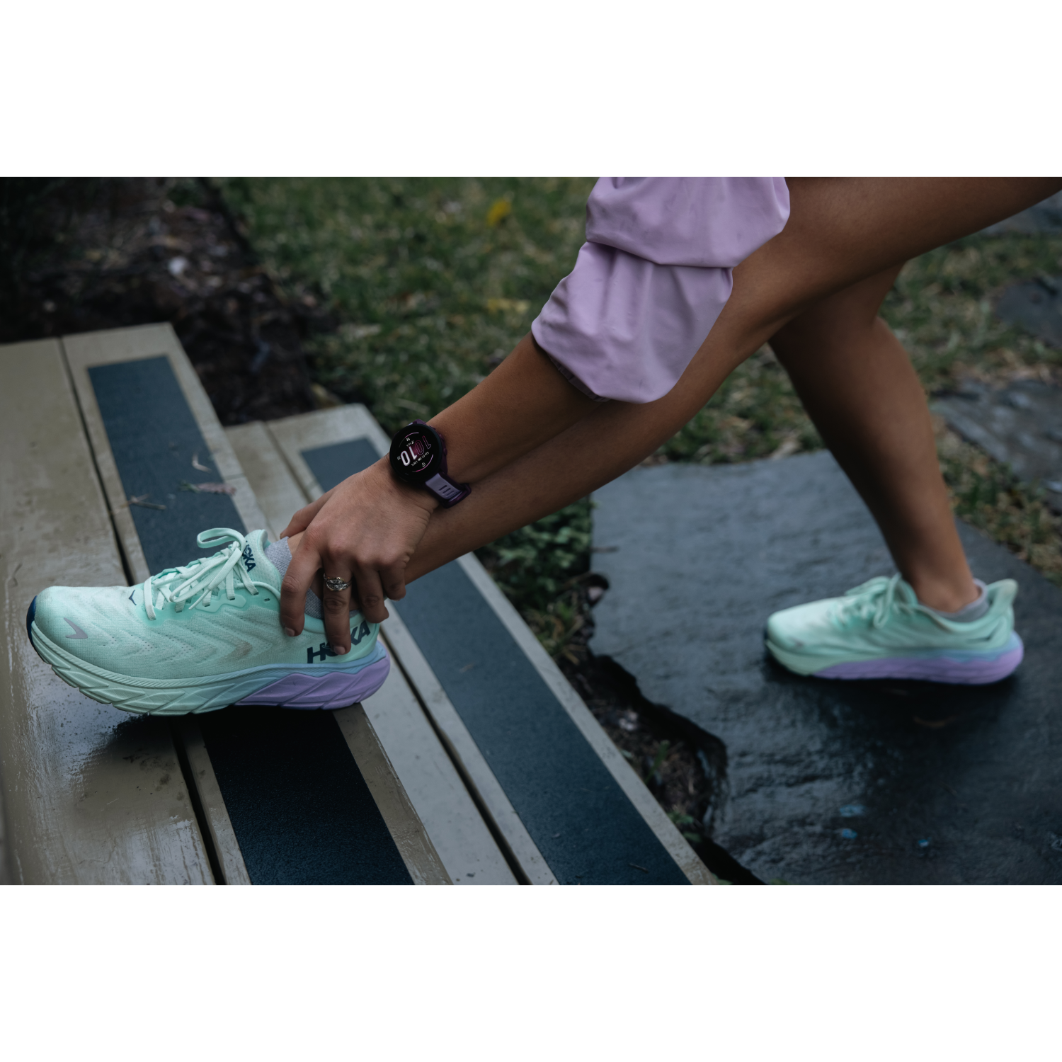 Person stretching leg with Hoka running shoe visible. Other running shoe is on the ground. Black watch on wrist.