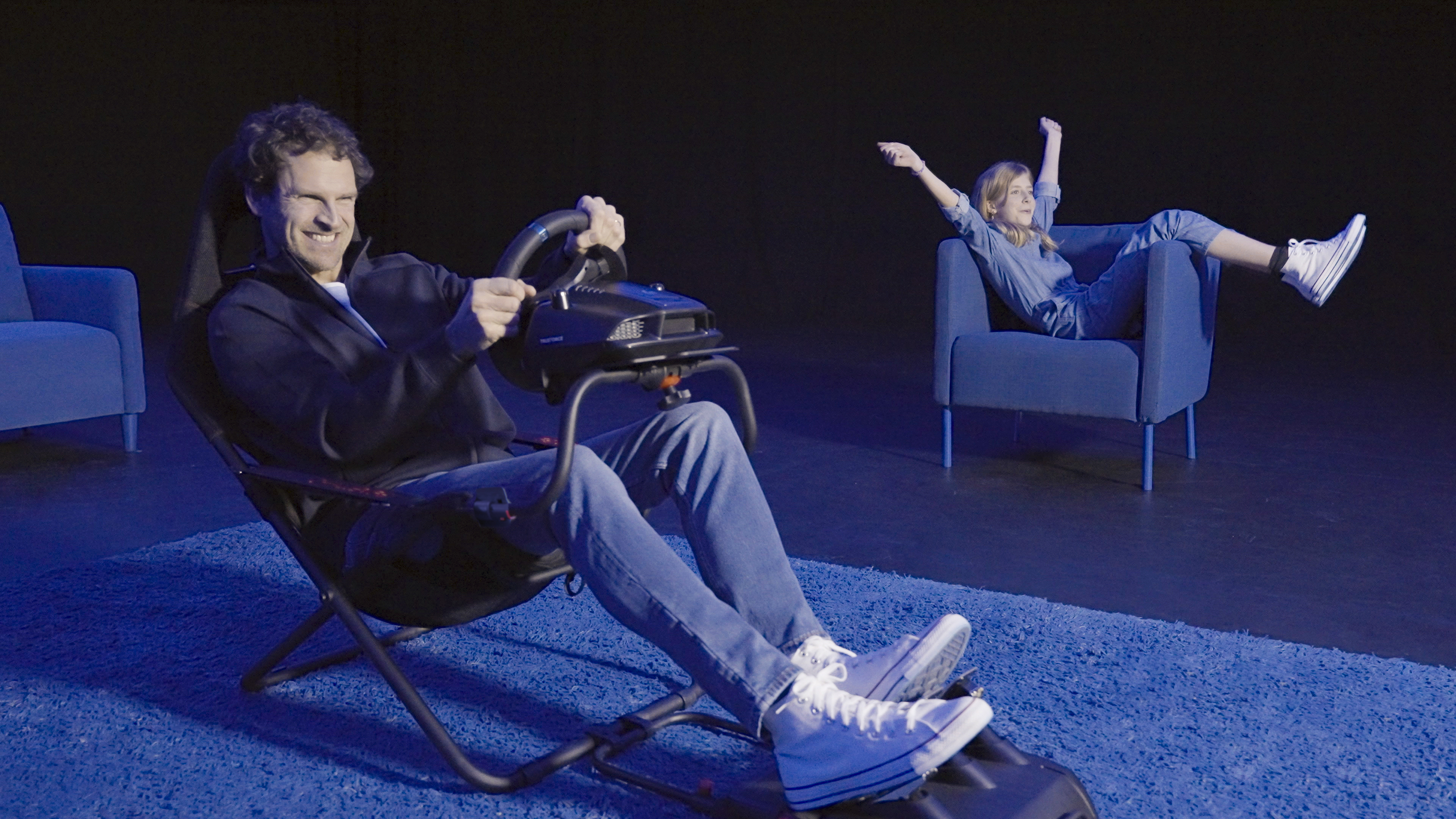 Man in racing seat holding steering wheel, girl relaxing in armchair.