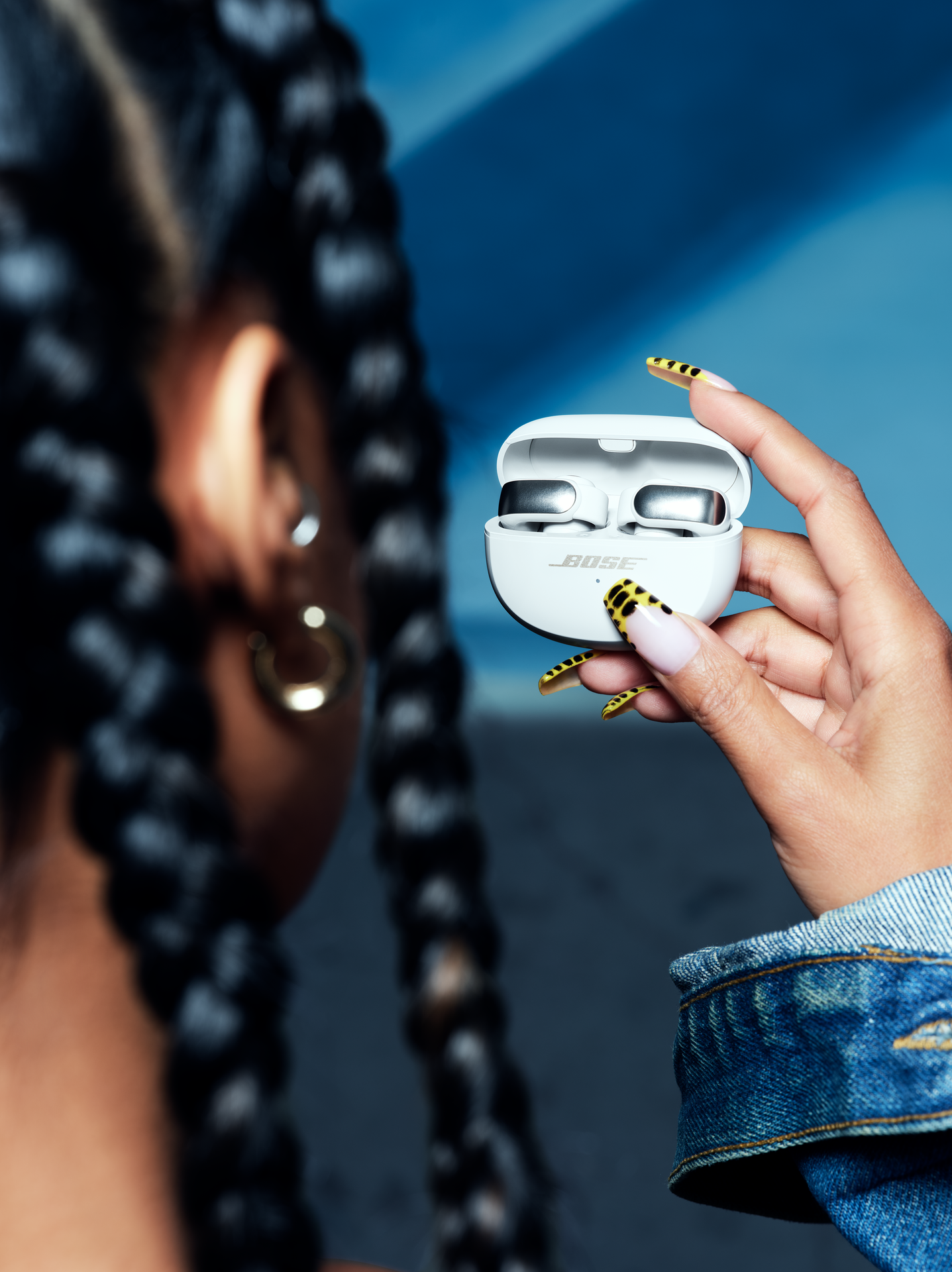 Kobieta trzyma białe etui na słuchawki Bose, w dżinsowej kurtce i z żółtym lakierem do paznokci.