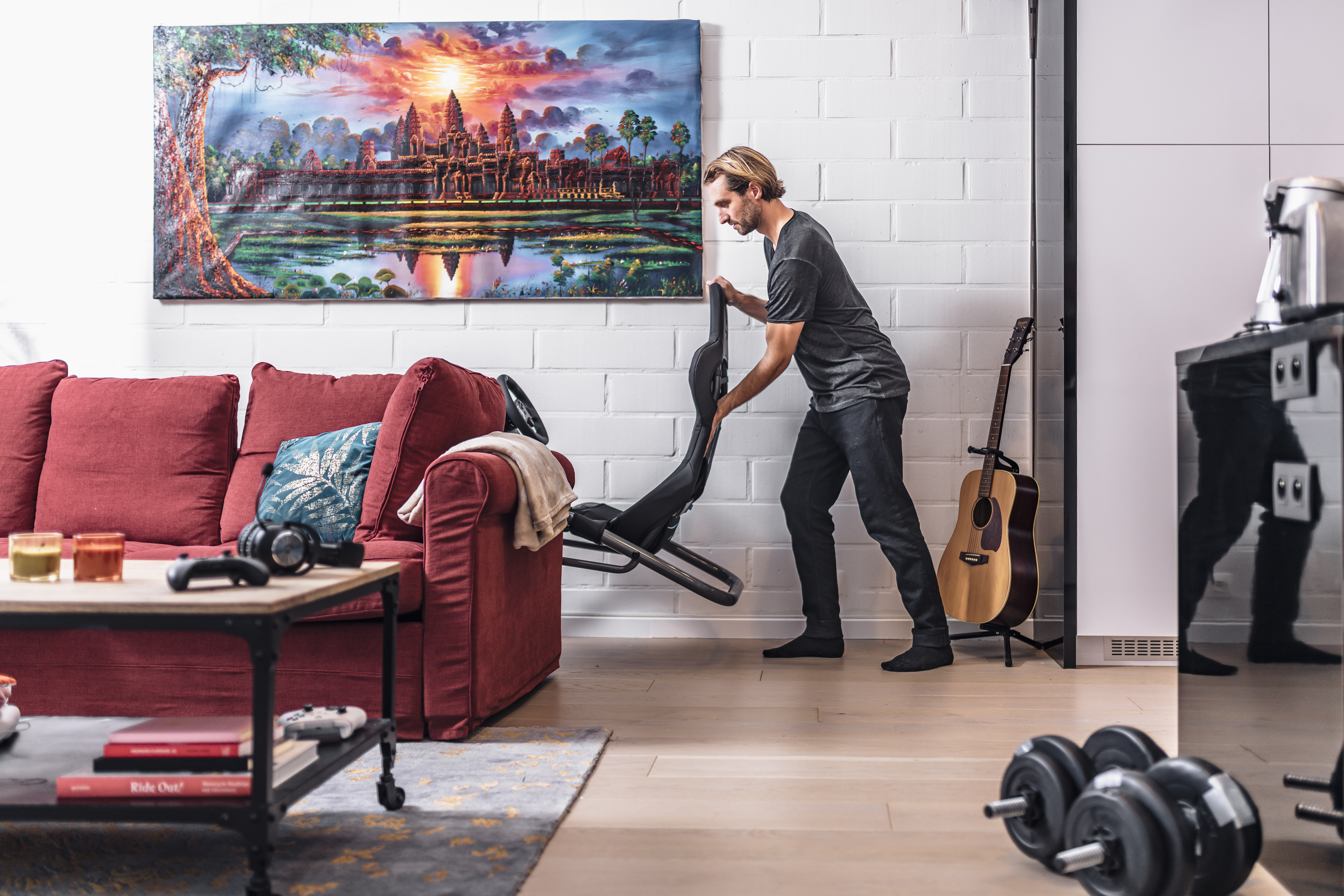 Un homme soulève une chaise de jeu noire dans un salon. Un canapé rouge, une guitare et une peinture d'un temple sont visibles.