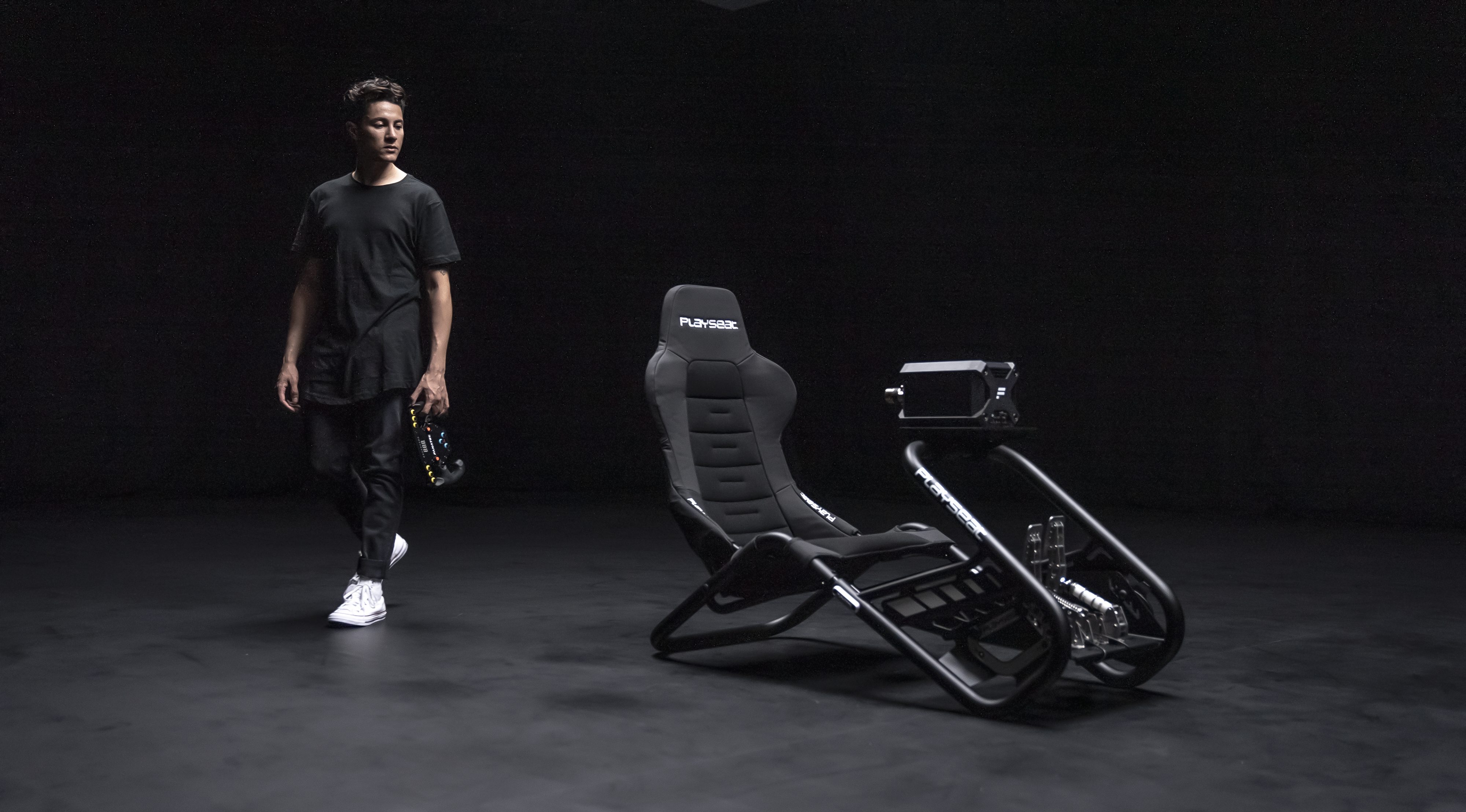 Un homme marche vers un siège de simulateur de course. La chaise est noire et grise, avec un volant. Le fond est sombre.