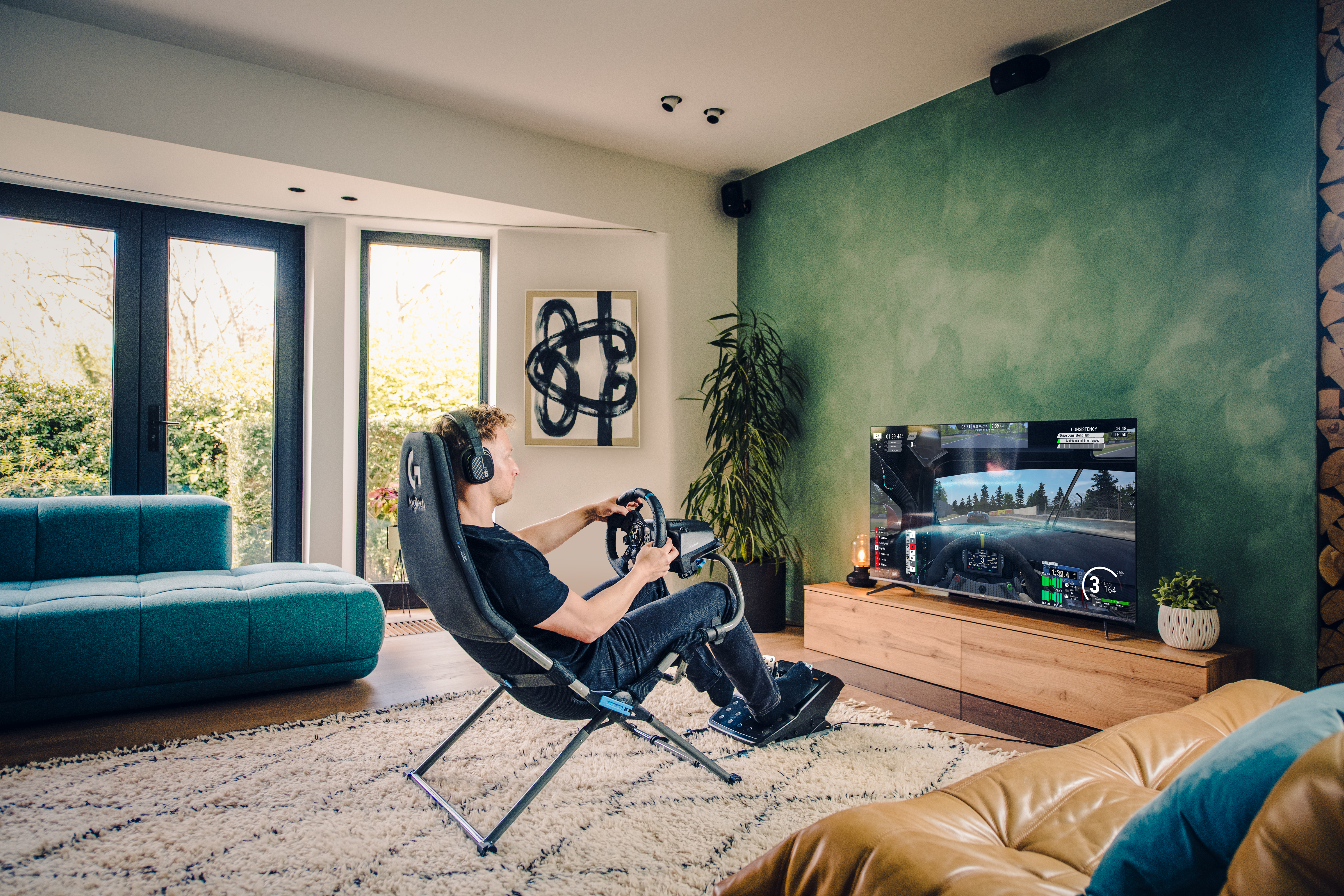 Man plays racing game with steering wheel and pedals in a living room.