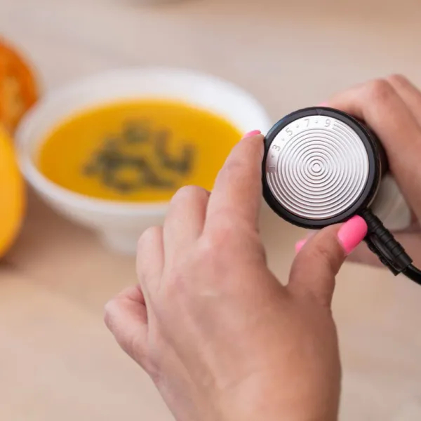 Dłonie kobiety z różowymi paznokciami regulują pokrętło urządzenia. W tle miska z zupą.