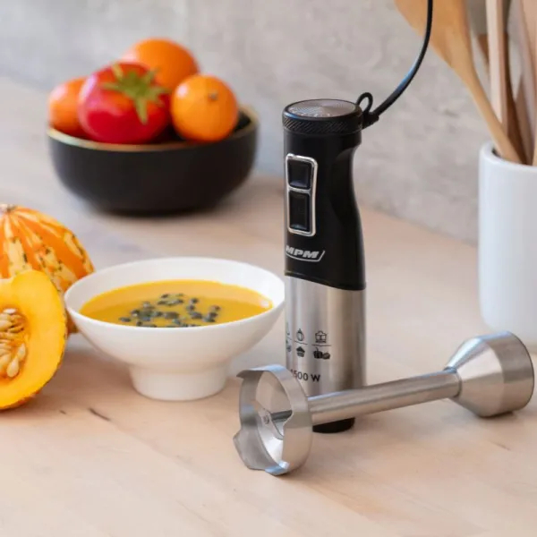 Blender ręczny i miska zupy na stole z owocami i drewnianymi łyżkami.