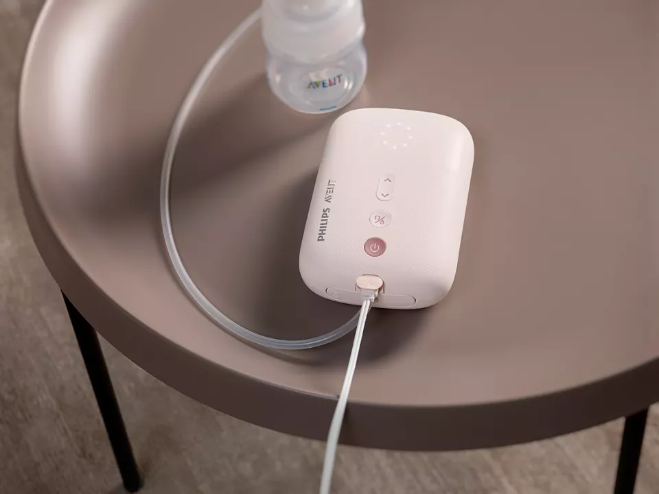 Egy fehér és rózsaszín Philips Avent mellszívó egy barna asztalon, a közelben egy cumisüveggel.