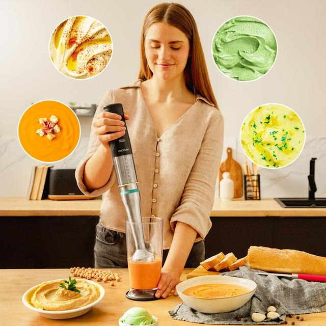 Mujer usando una batidora en una cocina. Hay cuencos con hummus, sopa y puré de patatas. La batidora está en un vaso de zumo.