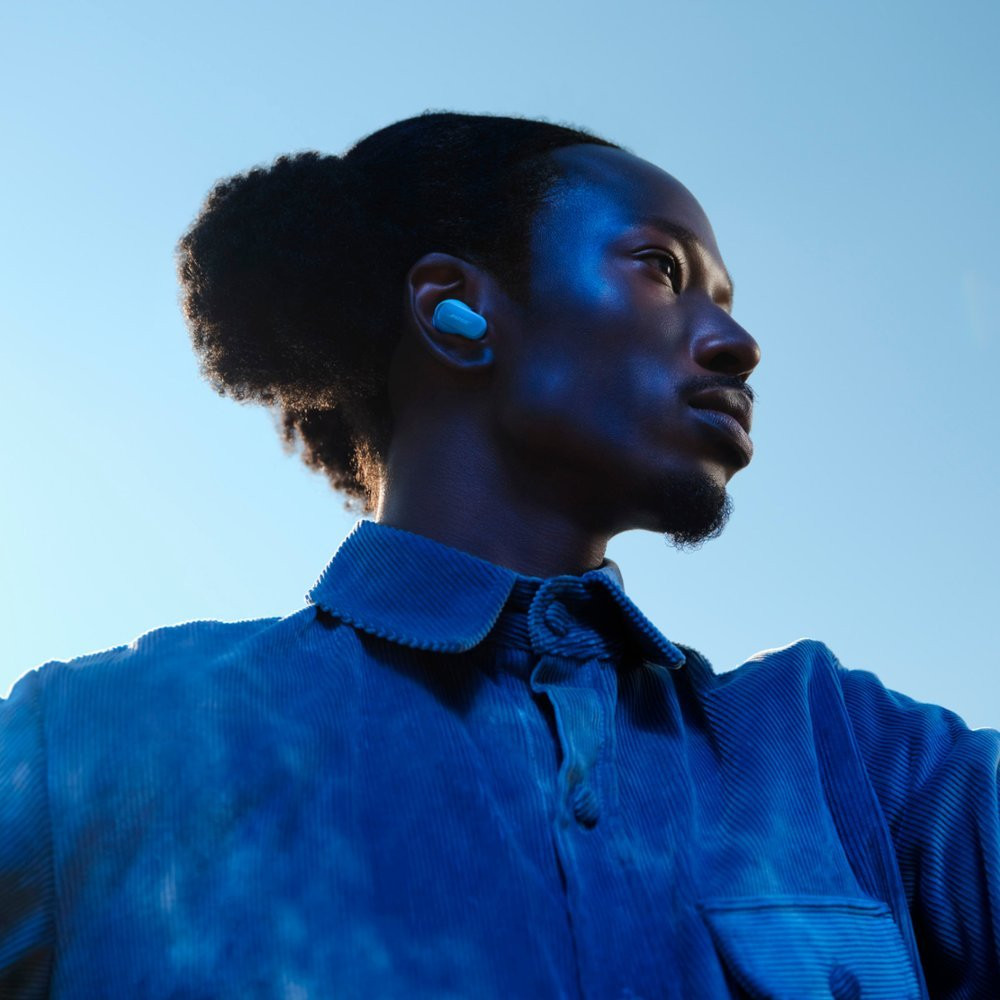 A man with blue light on his face, wearing a blue shirt, and one earbud in his ear.