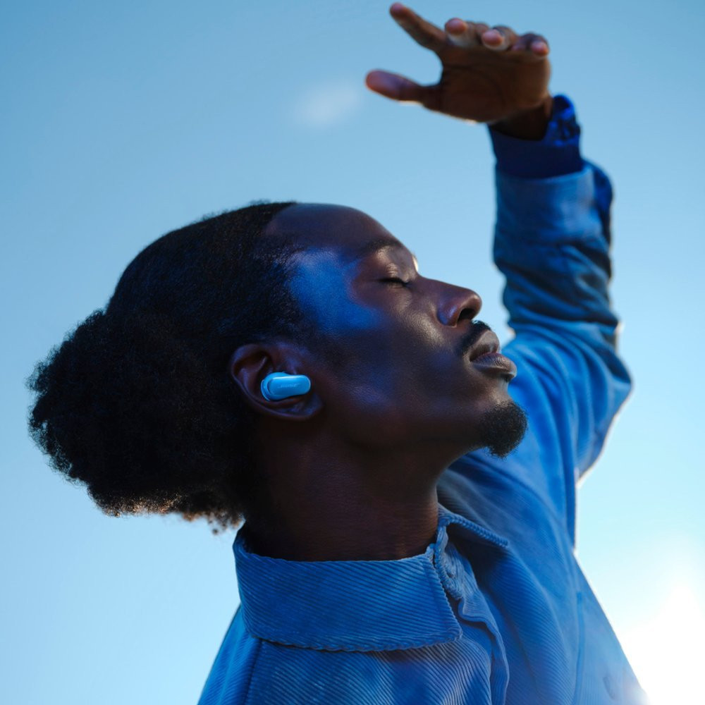 Man with blue earbud, looking up at bright sky, hand raised towards the sun.