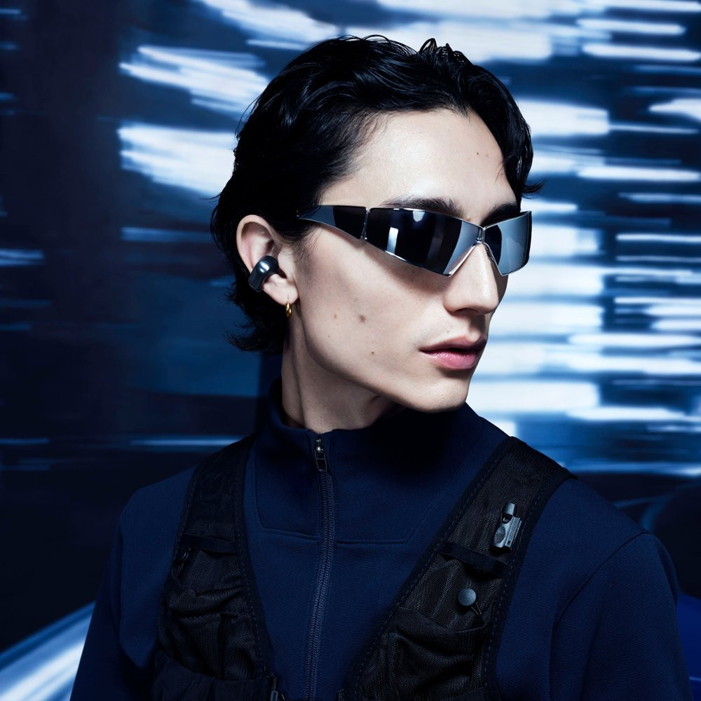 Young man wearing sunglasses, black vest, and earbud. Blue background. Modern, stylish appearance.