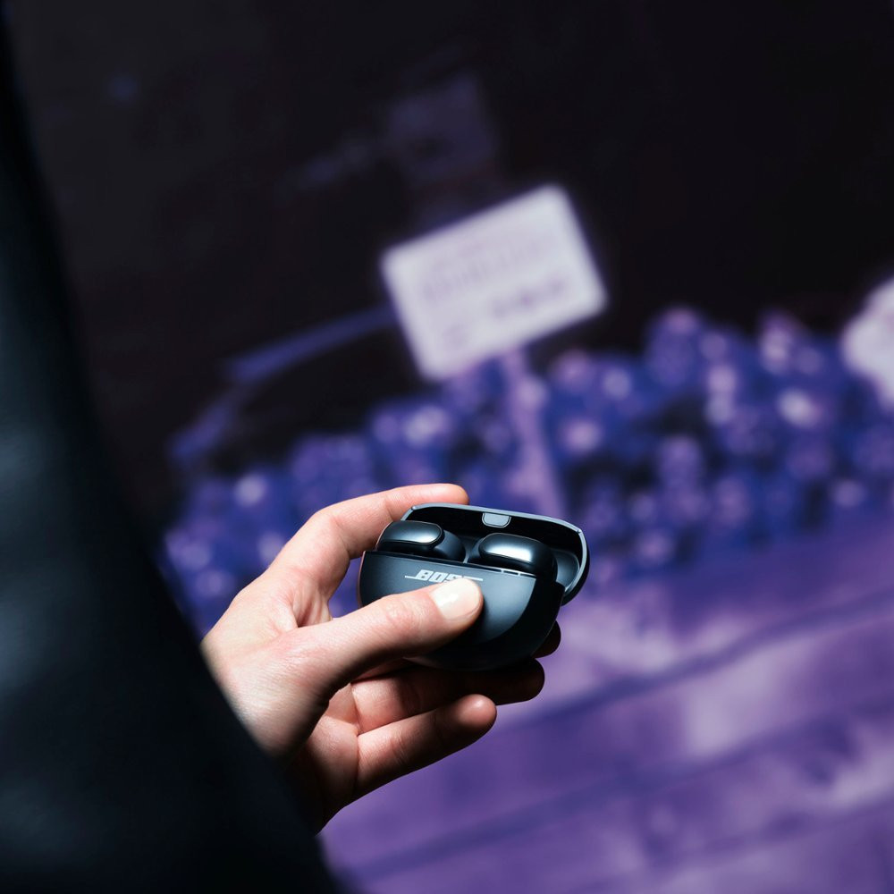 Hand holding black earbuds in an open charging case, with a blurred background of a shop.