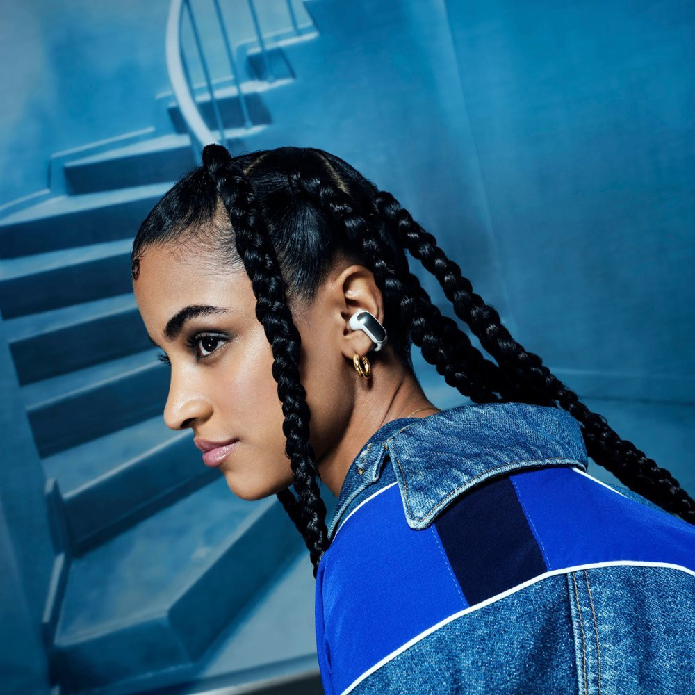 A woman wearing a white and black earbud, with braids and a blue jacket.