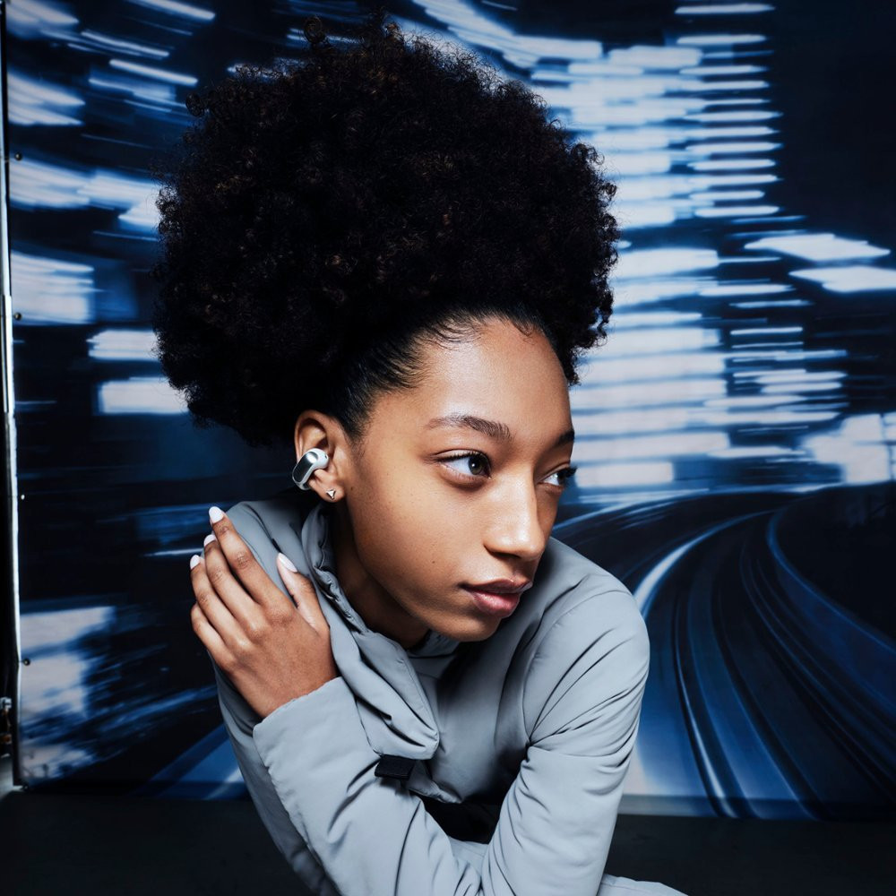 A young woman wears earbuds. She has dark curly hair and looks to her right. A blue blurred background.