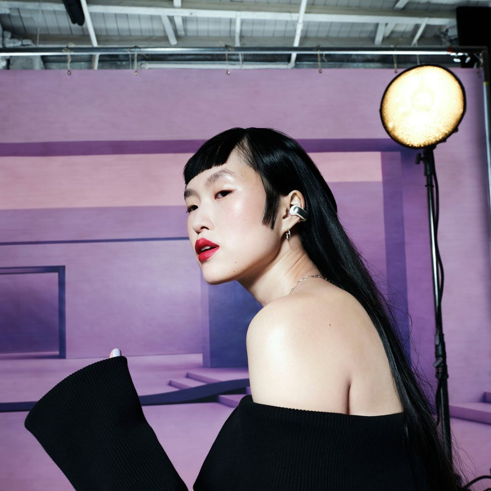 Asian woman in black outfit and earrings, red lipstick, looking to the side. Studio setting.